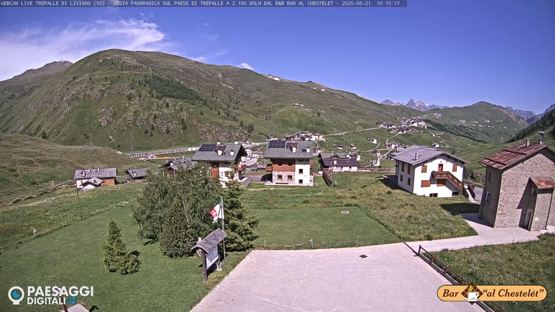 A mountain village nestled in a valley on a sunny day, with green hills and mountains in the background.