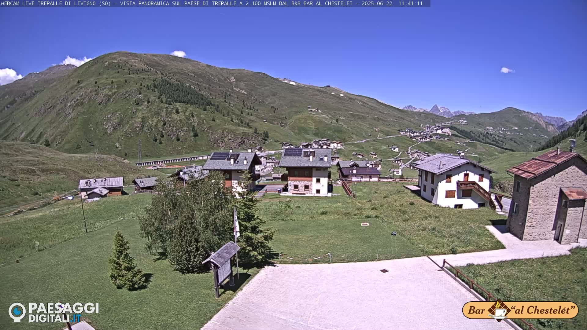A sunny mountain village nestled in a valley, surrounded by green hills and mountains under a clear blue sky.