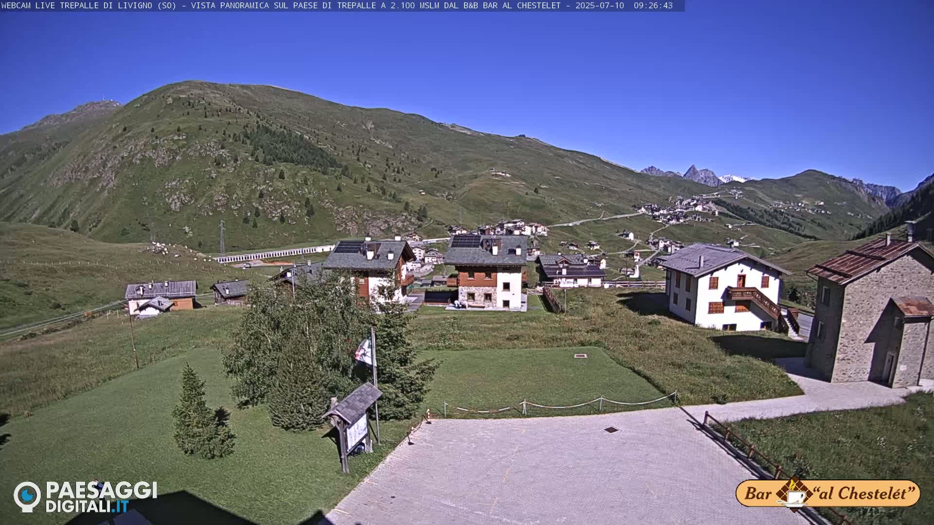 A mountain village with several buildings is nestled in a valley on a sunny day, surrounded by rolling green hills and distant snow-capped peaks.