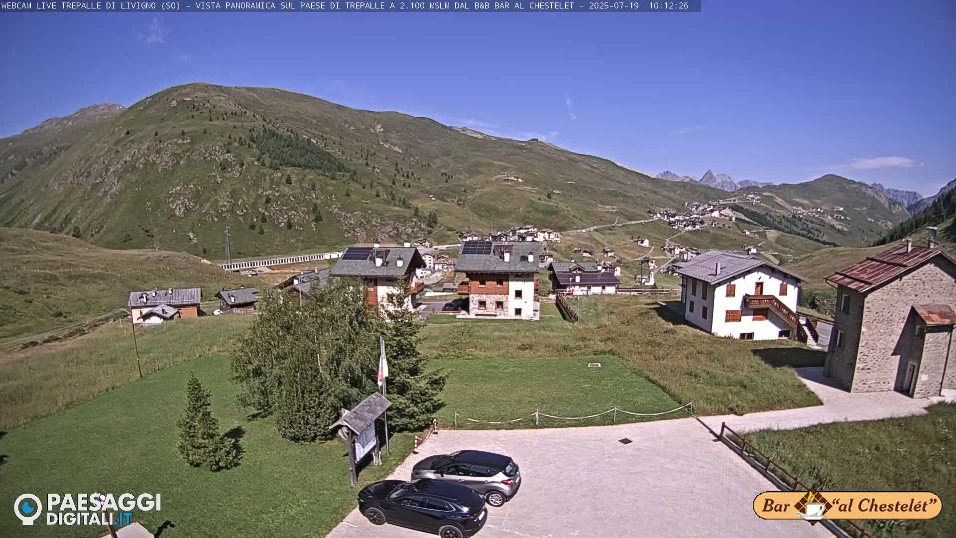 A mountain village with several buildings, two cars parked in a lot, and green grass, all under a clear blue sky.