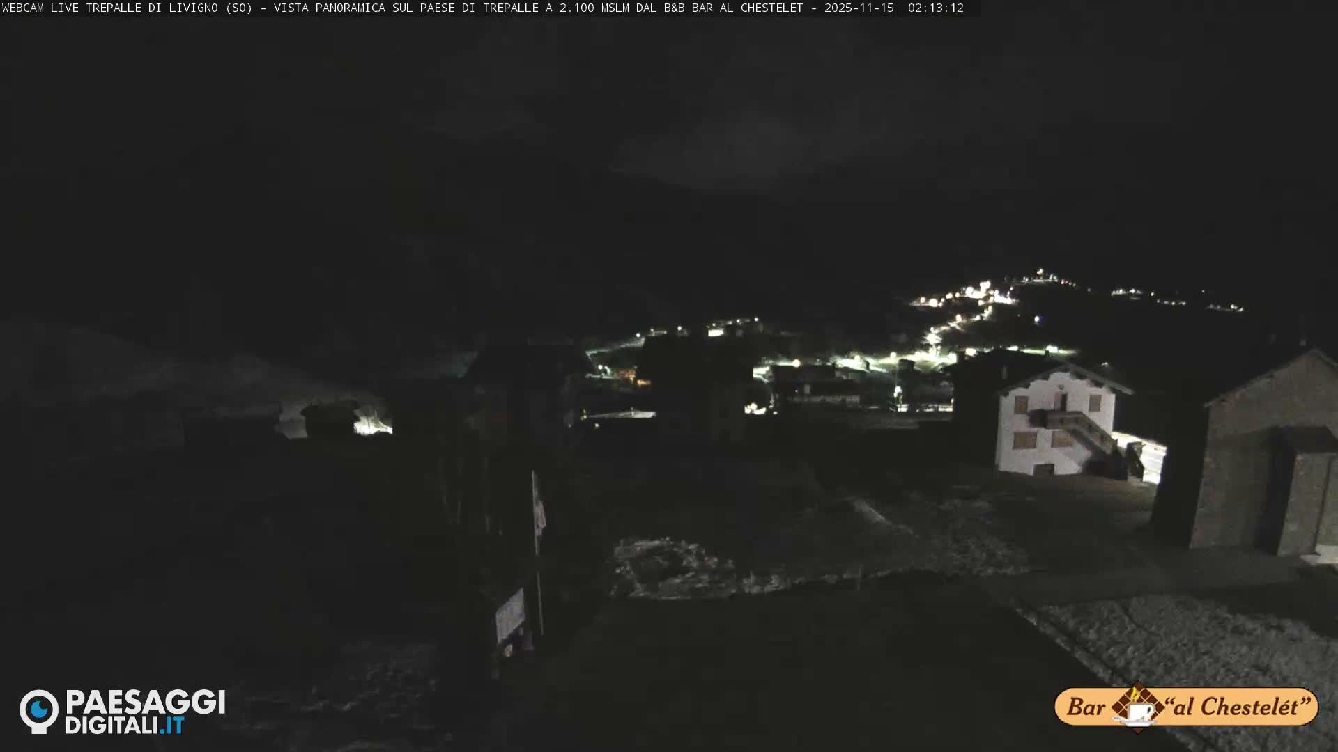 A nighttime view of a snow-dusted mountain village under a clear sky, with scattered lights illuminating buildings and roads across the hilly terrain.