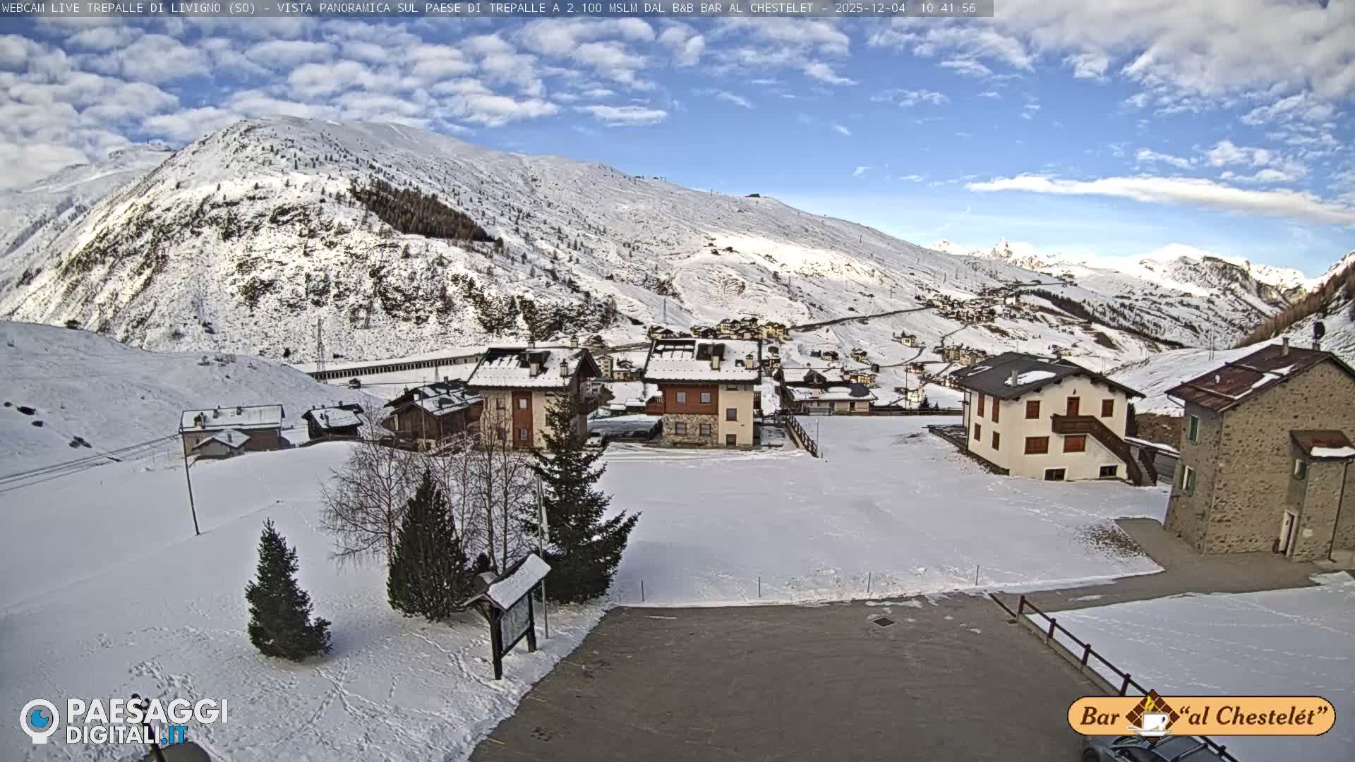A wide view displays a snow-covered mountain village featuring multiple houses and evergreen trees, with vast snowy peaks rising in the background under a blue sky dotted with white clouds, indicating clear and cold weather.