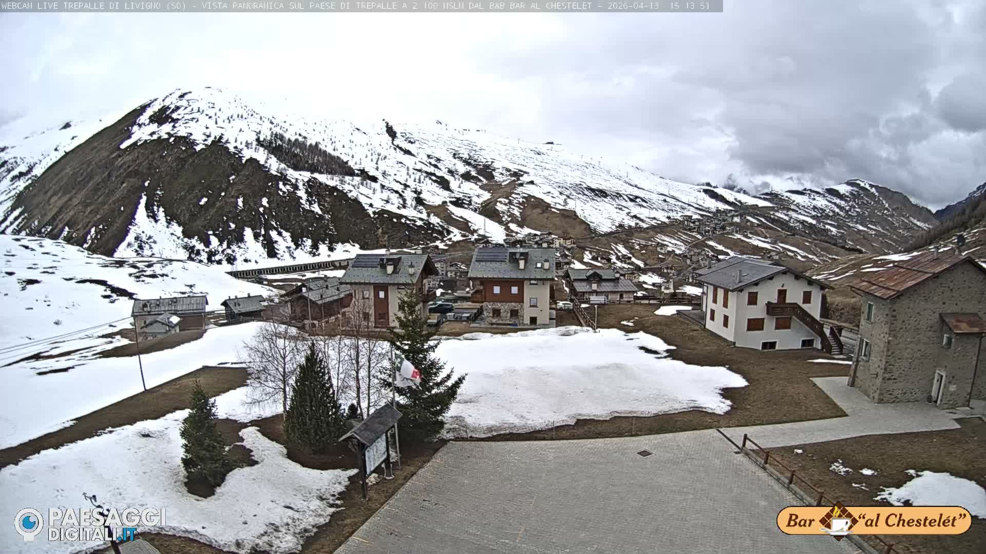 A wide view displays a snow-covered mountain village featuring multiple houses and evergreen trees, with vast snowy peaks rising in the background under a blue sky dotted with white clouds, indicating clear and cold weather.