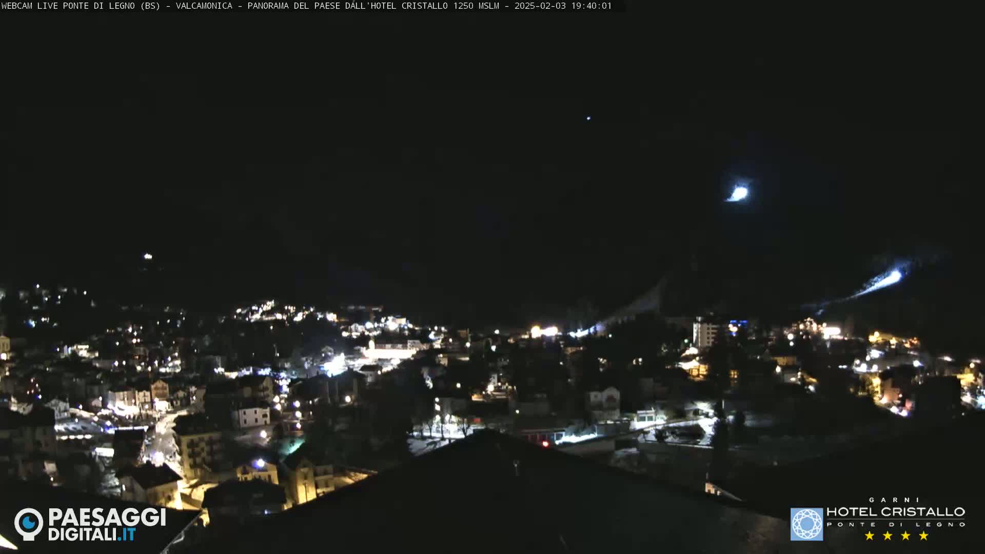 A nighttime view of a snow-covered town illuminated by lights, with two bright streaks visible in the dark sky above.