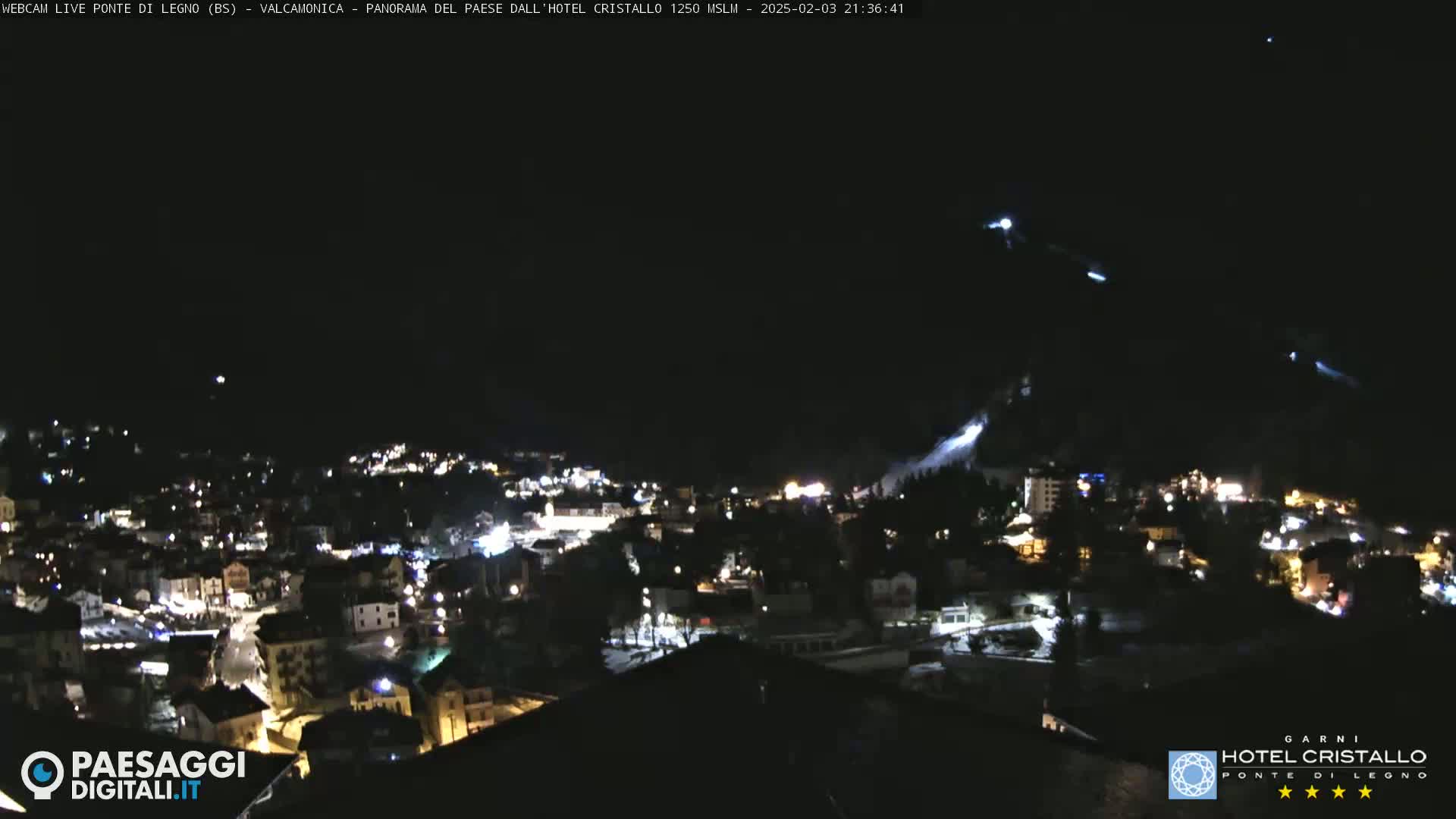 A nighttime view of a snow-covered town illuminated by lights, with streaks of what appears to be snow or ice falling from a nearby mountain.