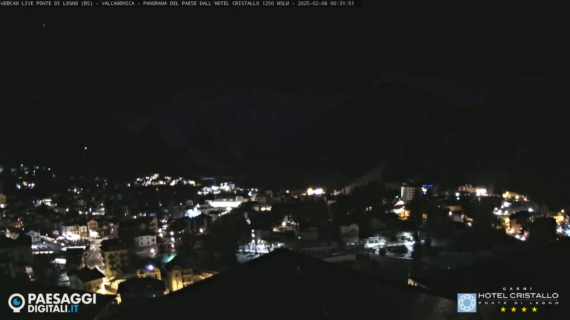 A nighttime, long shot of a town nestled in a valley, illuminated by scattered lights under a dark sky.