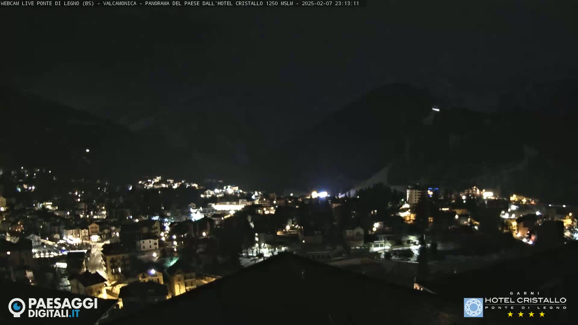 A nighttime view of a town nestled in a valley, illuminated by scattered lights, with dark mountains in the background under a clear, dark sky.