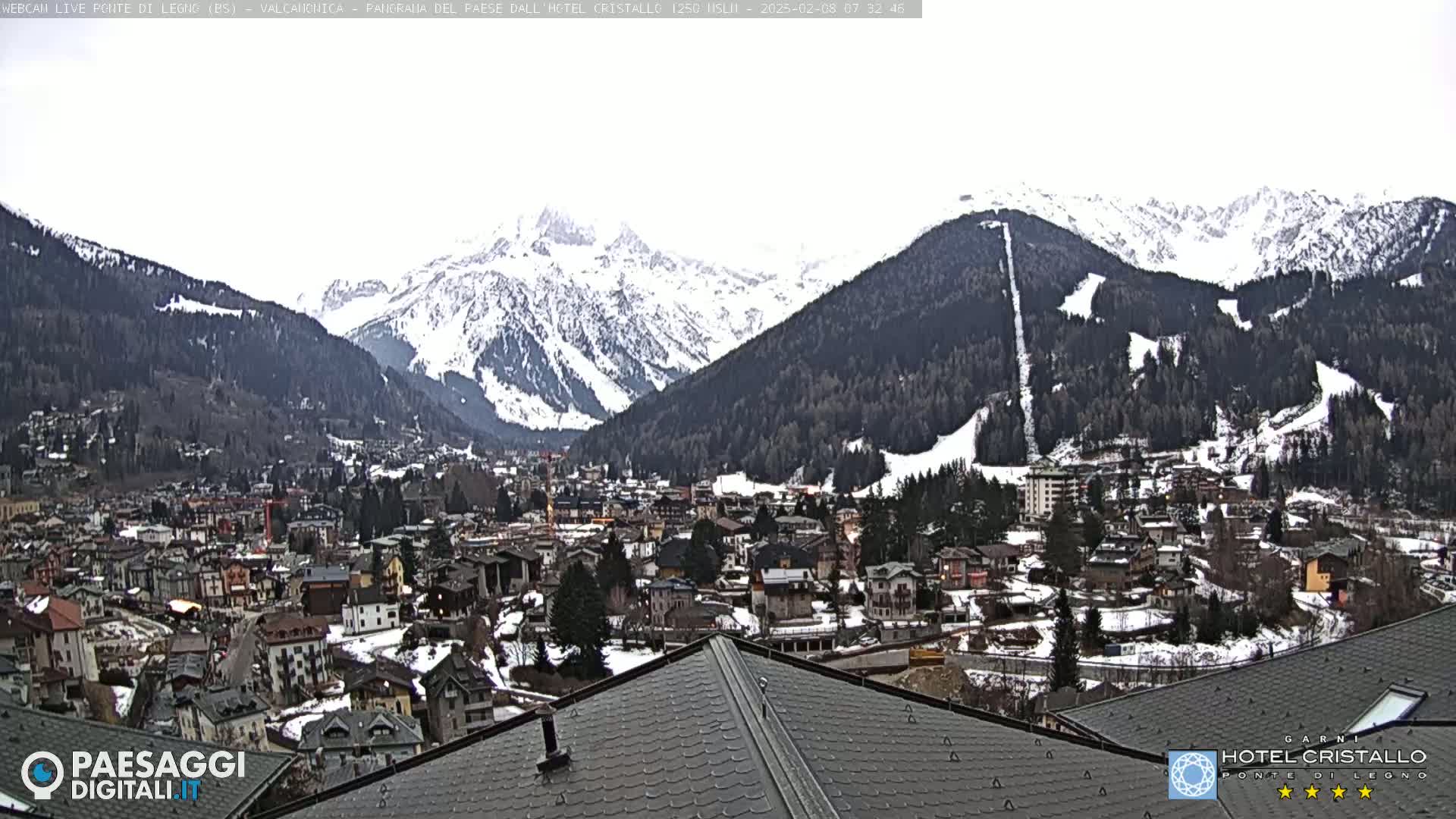 A snow-dusted mountain village nestled in a valley, with snow-covered mountains rising in the background under an overcast sky.