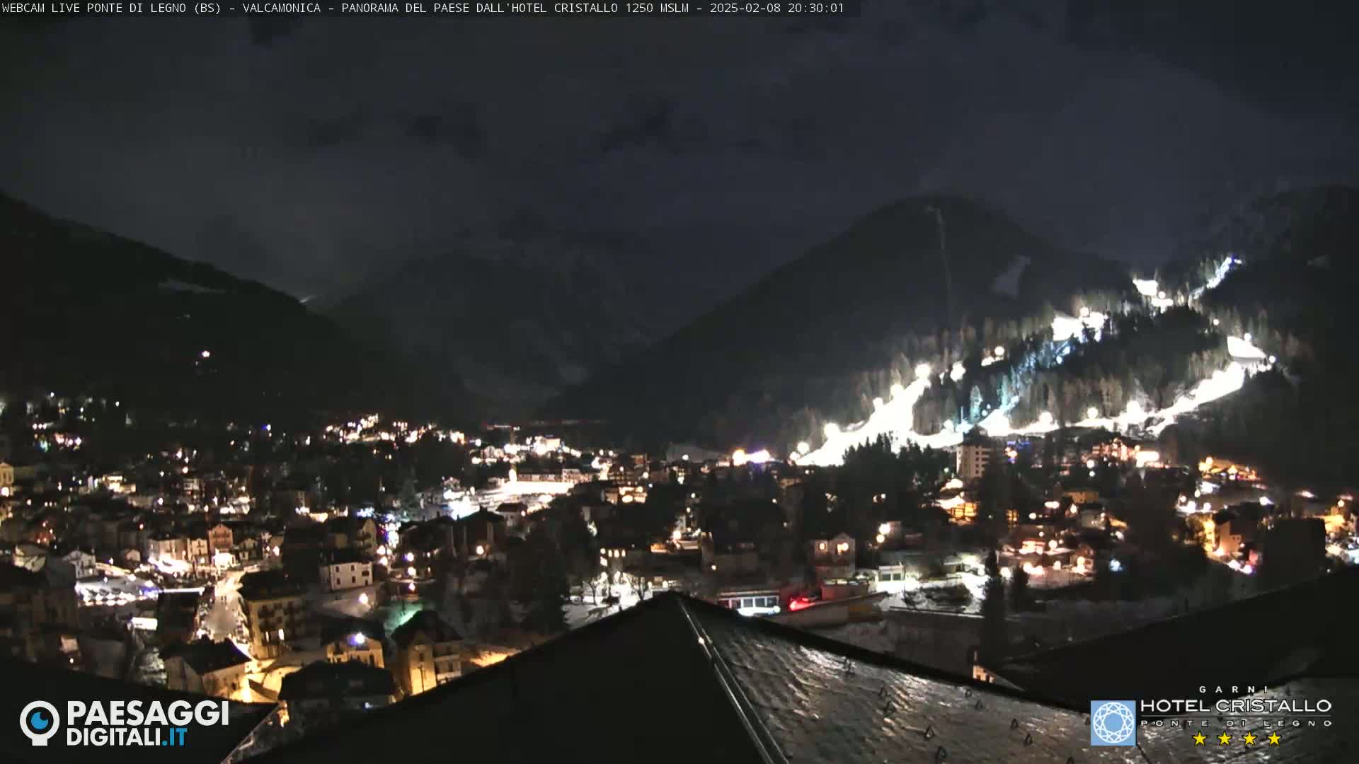 A nighttime view of a snow-covered mountain town illuminated by lights, with a dark, cloudy sky.