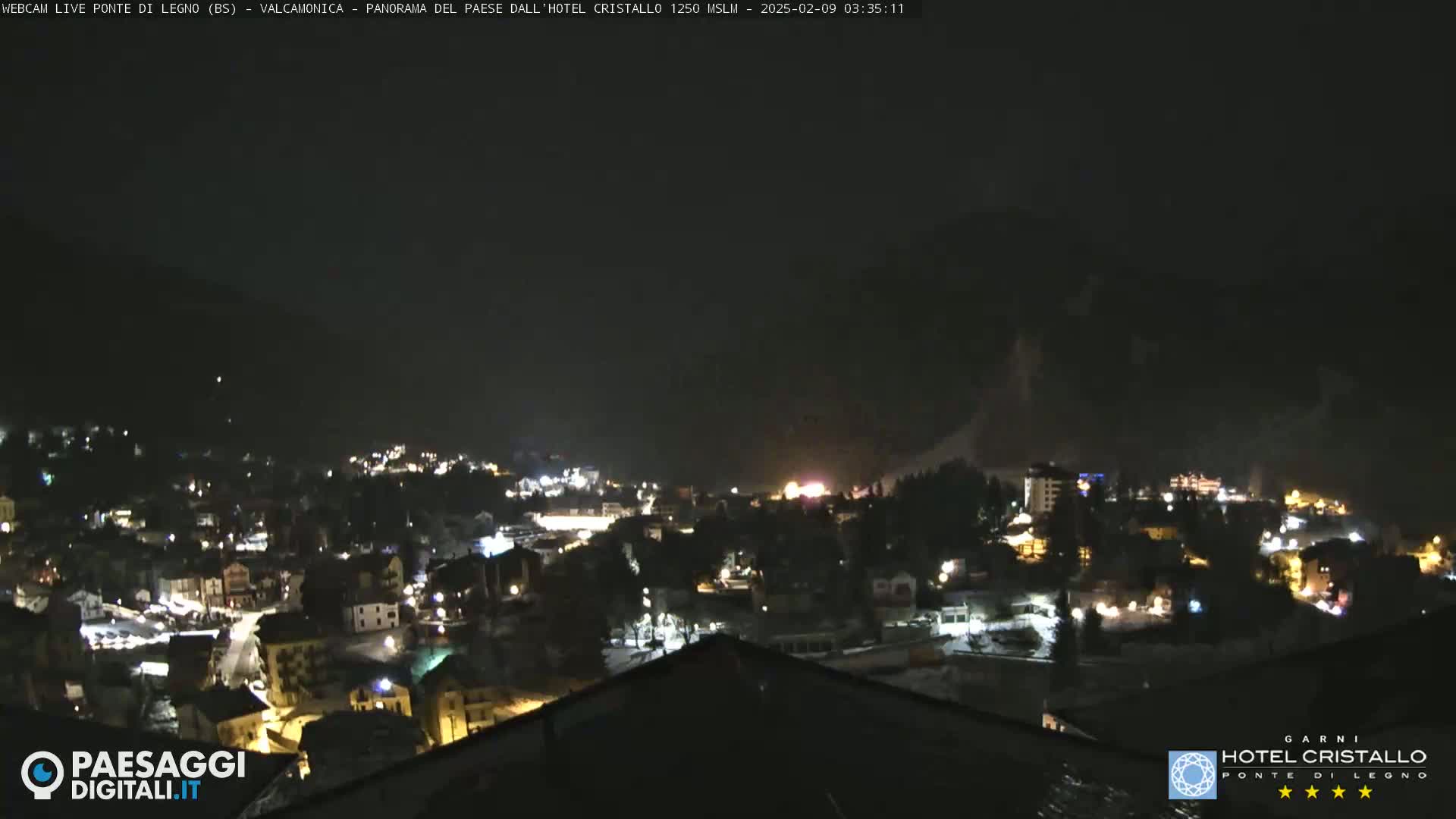 A nighttime view of a snow-covered town nestled in a valley, illuminated by scattered lights under a dark sky.