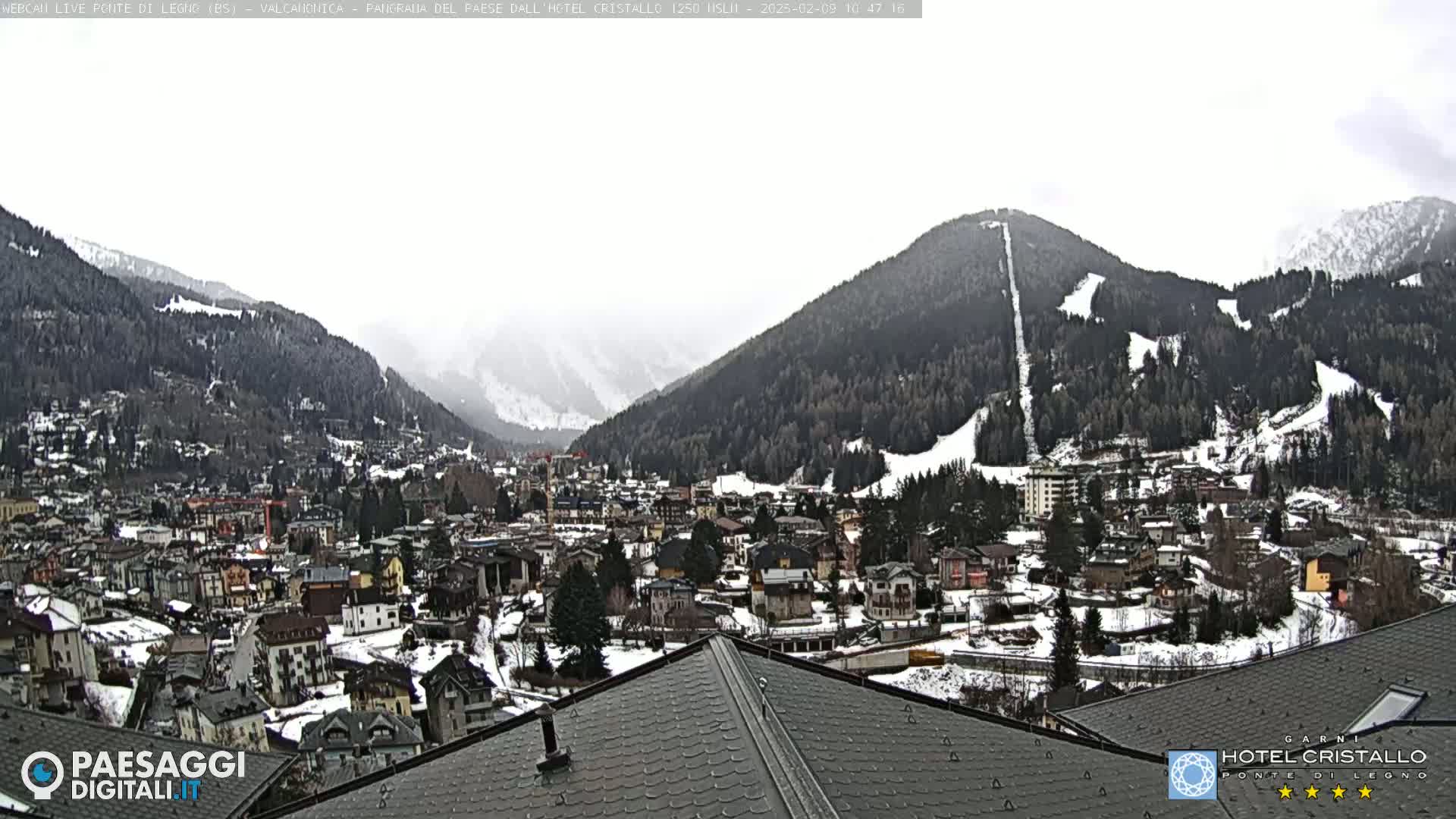 A snow-dusted mountain village nestled in a valley, with overcast skies and snow-covered slopes visible on surrounding mountains.