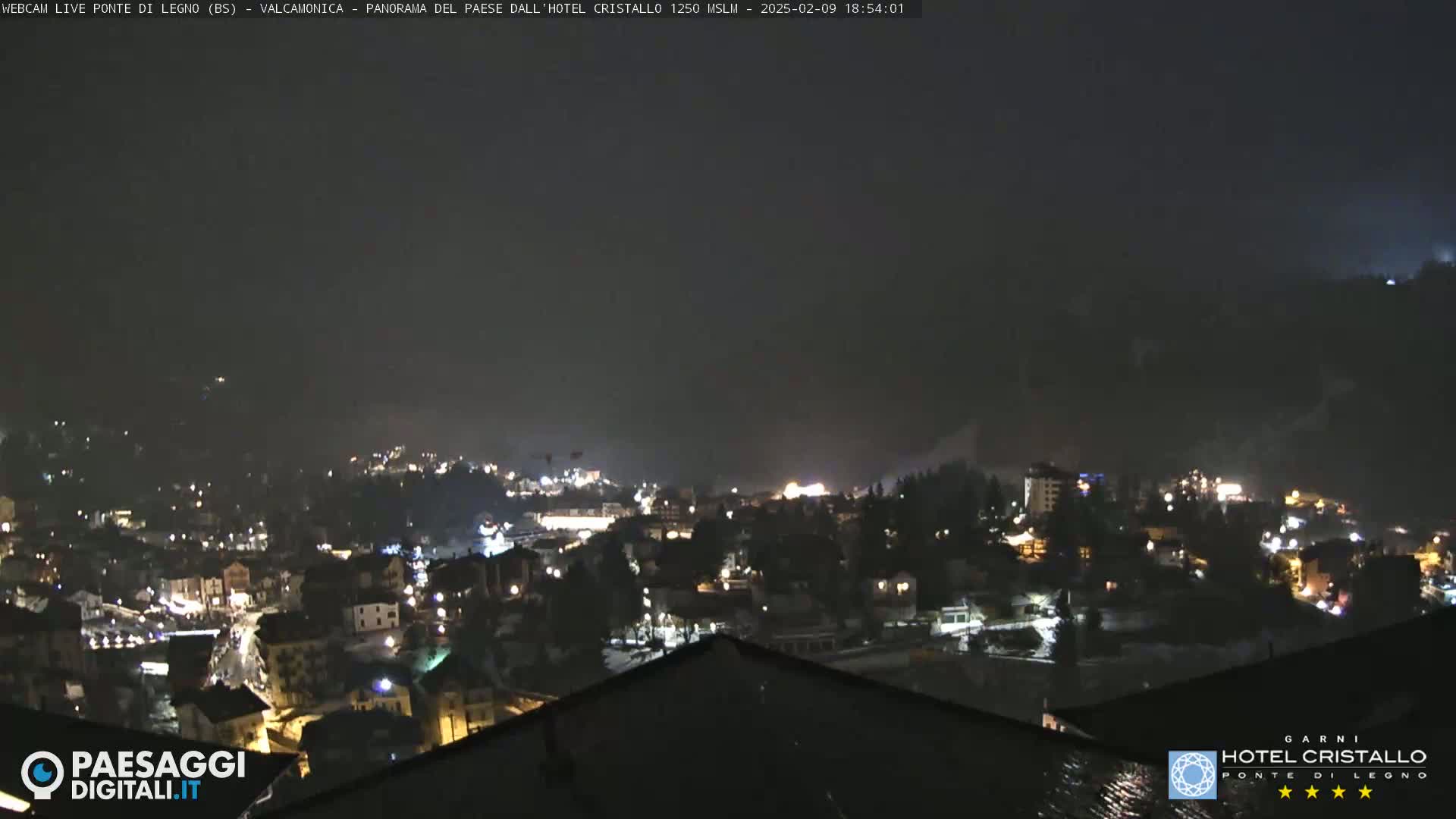 A nighttime view of a snow-dusted town nestled in a valley, illuminated by scattered lights under a dark, cloudy sky.