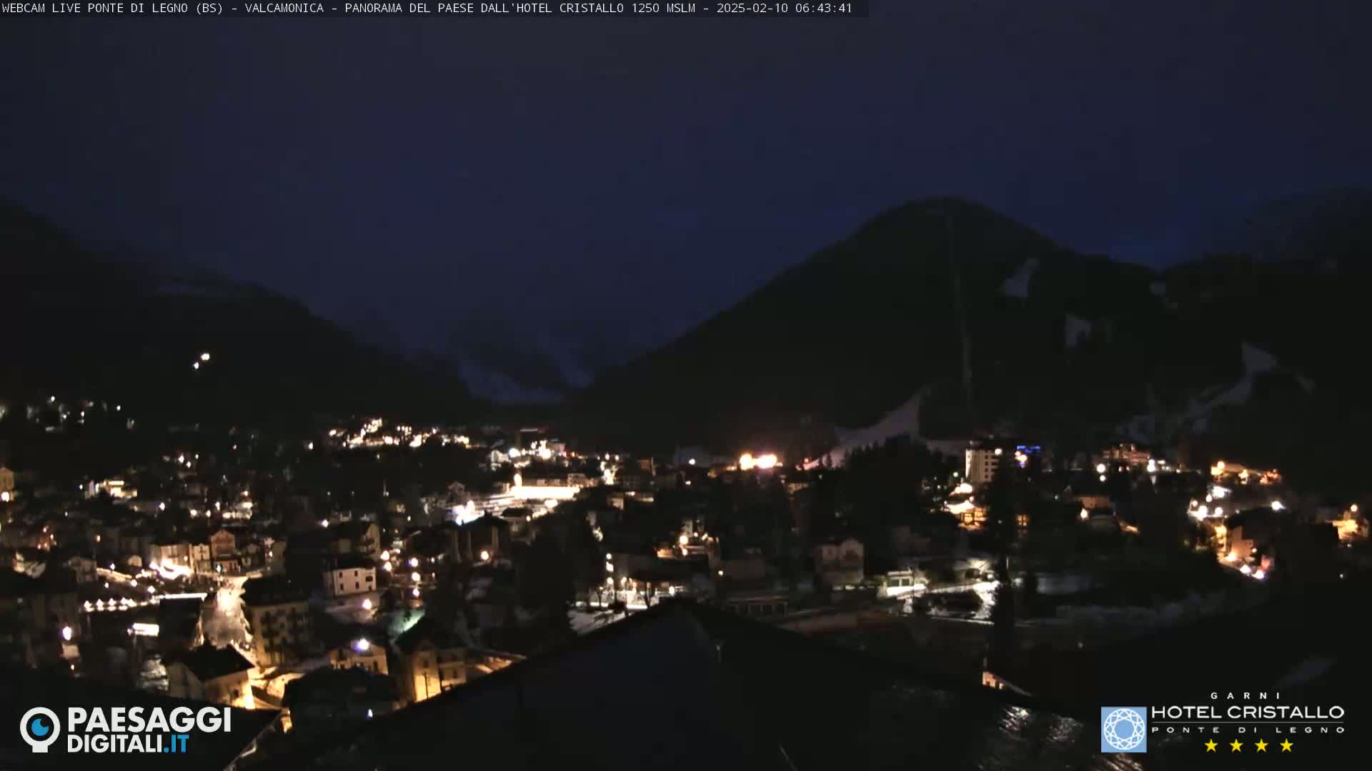 A nighttime, high-angle view shows a town nestled in a valley between dark mountains, illuminated by numerous streetlights.
