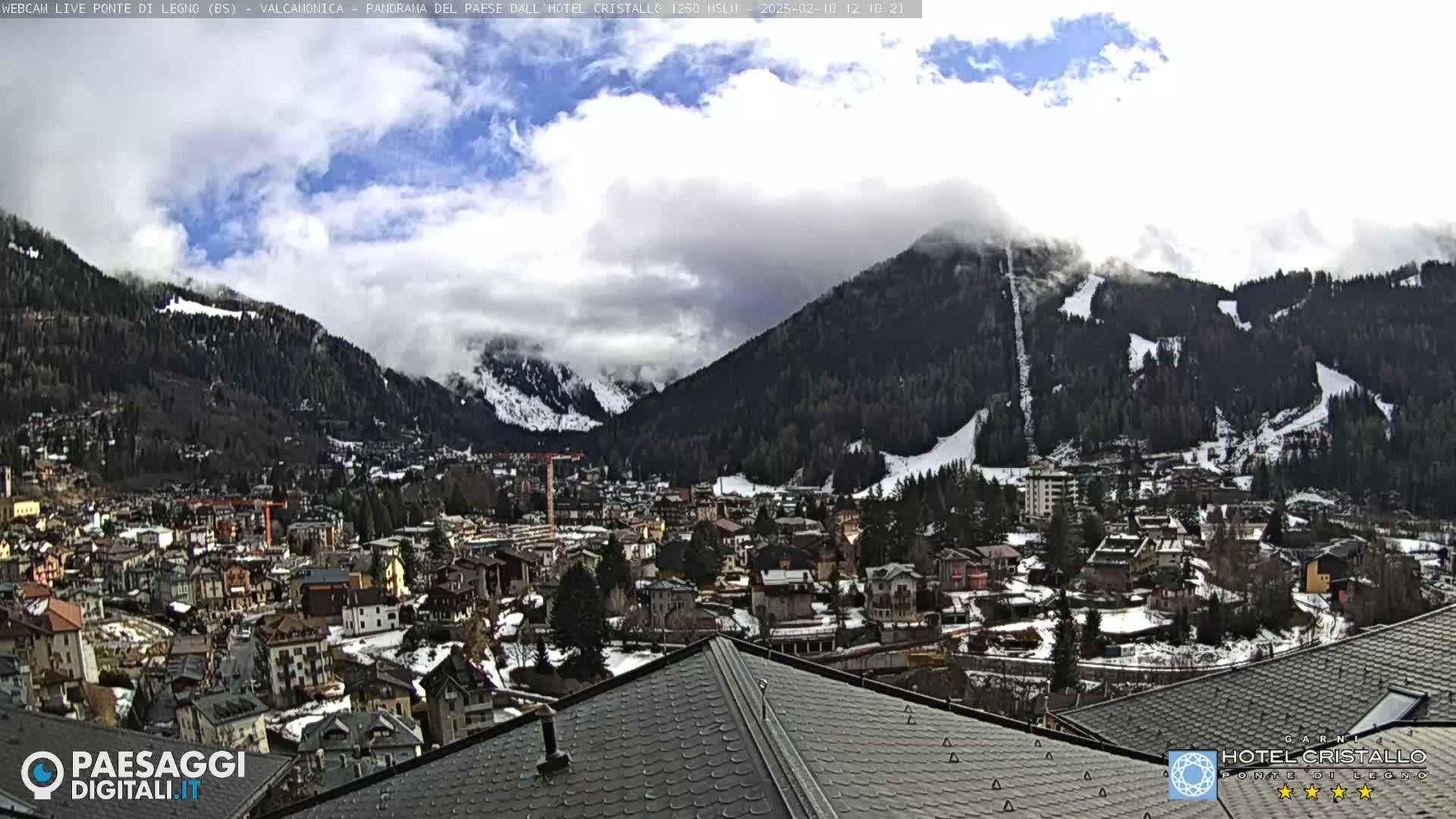 A partially cloudy day reveals a mountainous town with snow-dusted roofs and ski slopes, viewed from a rooftop.