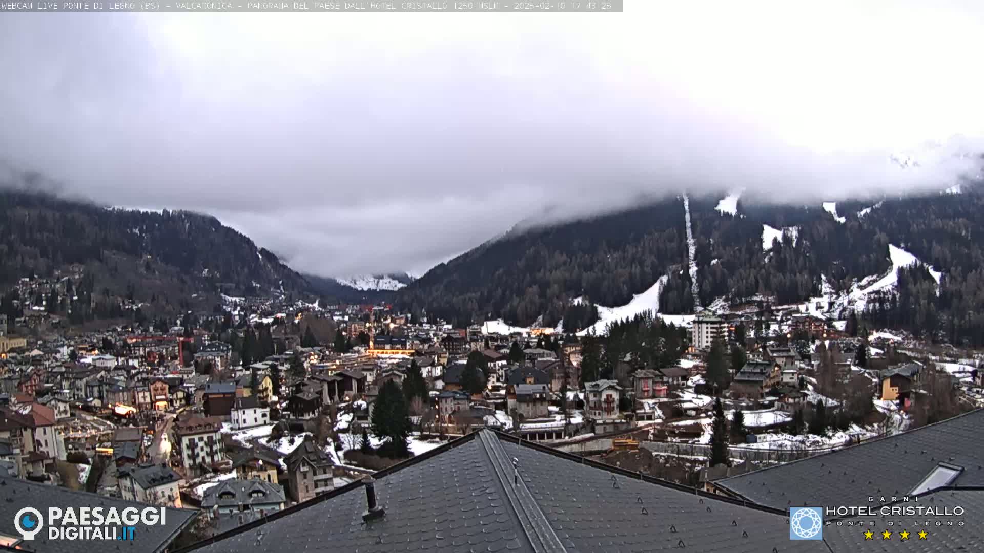 A partially cloudy, overcast view of a snow-dusted mountain town nestled in a valley, with ski slopes visible on the surrounding hills.