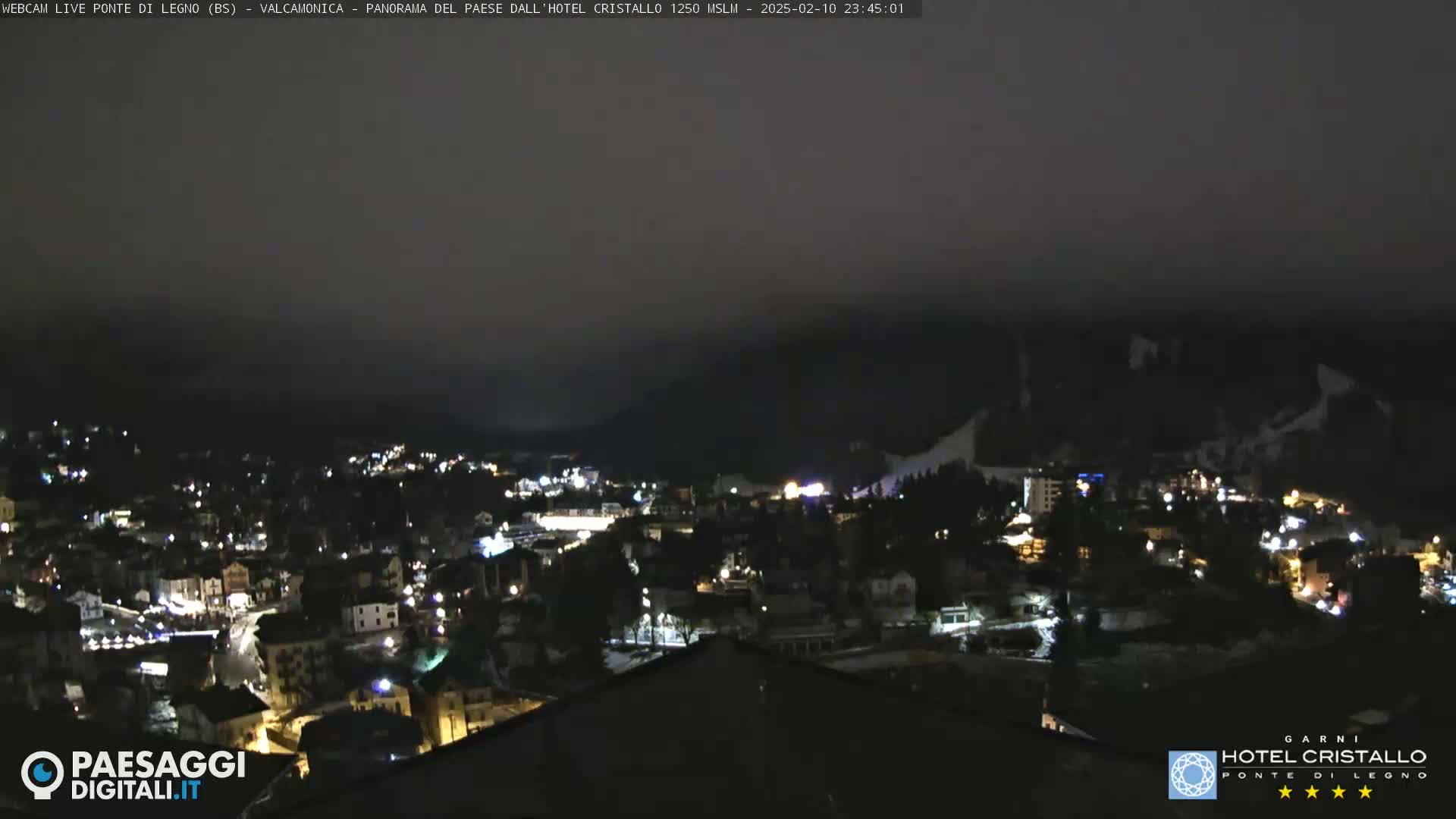A nighttime, overcast view of a snow-covered town nestled in a mountain valley, illuminated by scattered lights.