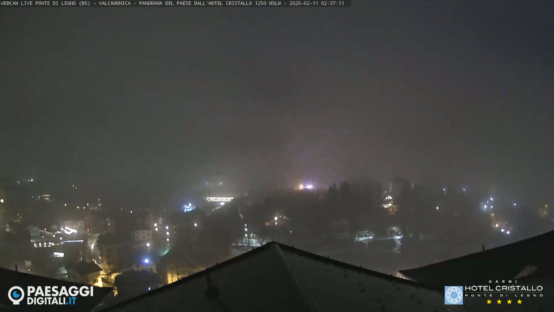 A nighttime, foggy view of a town nestled in a valley, with scattered lights visible through the haze.