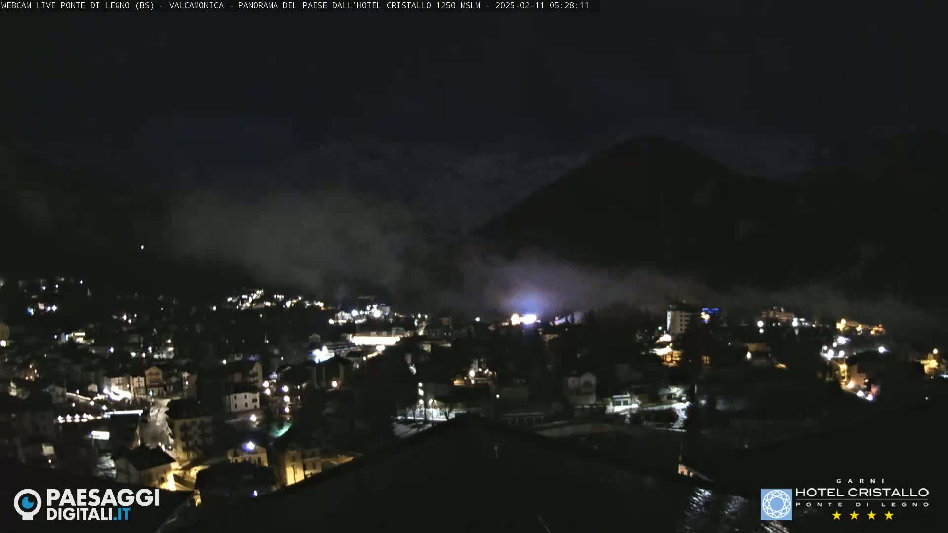 A nighttime view of a town nestled in a valley, partially obscured by fog or low clouds, with mountains in the background.