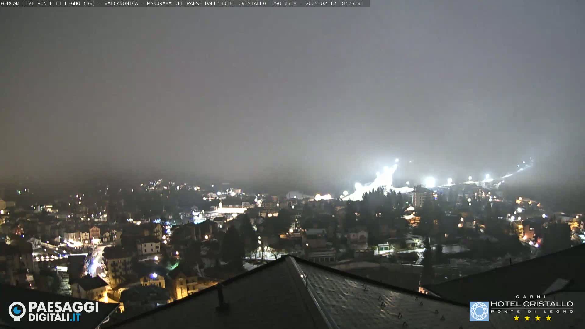 A nighttime, hazy view of a town illuminated by lights, with a brightly lit ski slope visible in the distance.