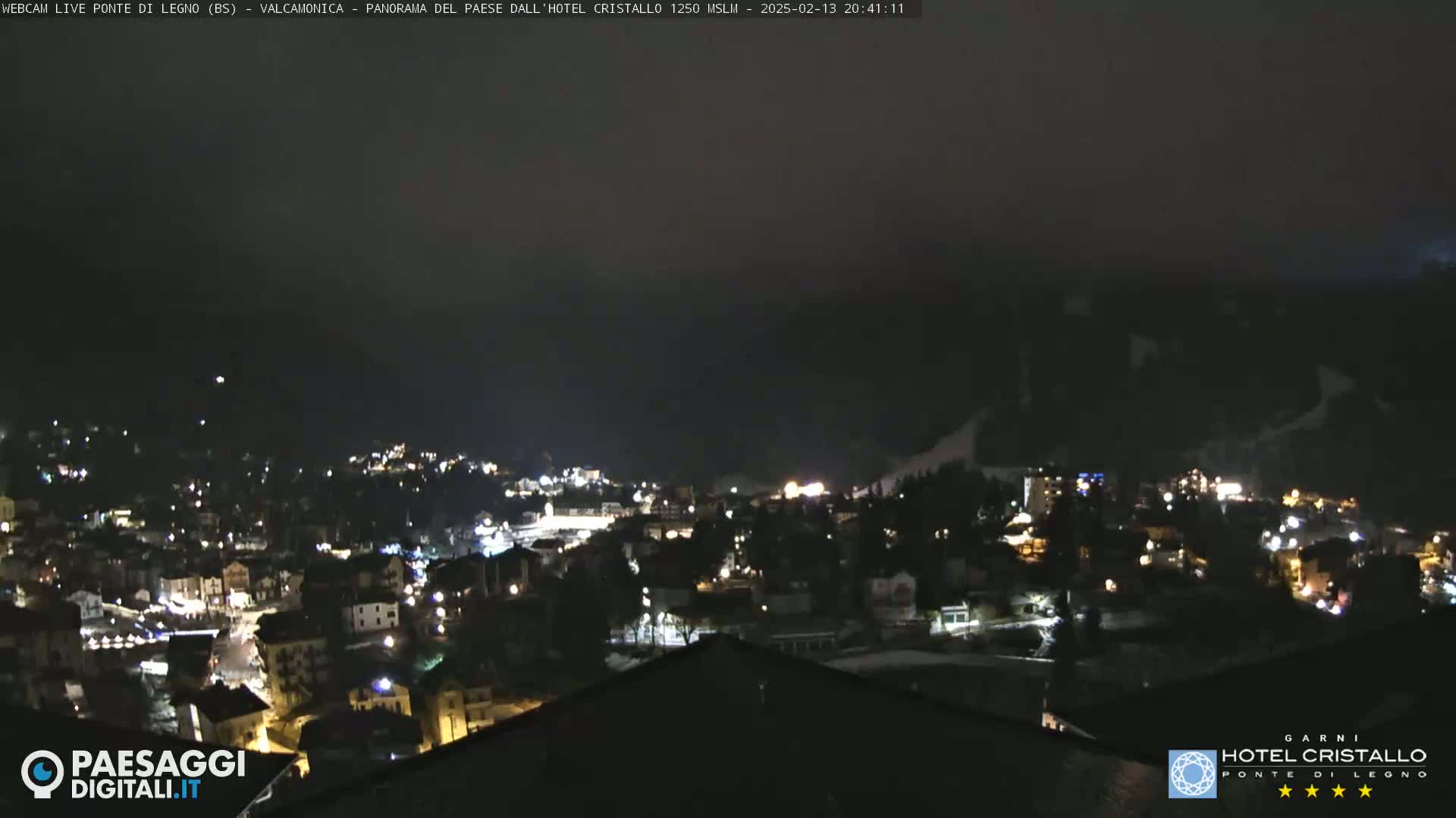 A nighttime, hazy view of a town illuminated by lights, nestled in a valley surrounded by dark mountains.