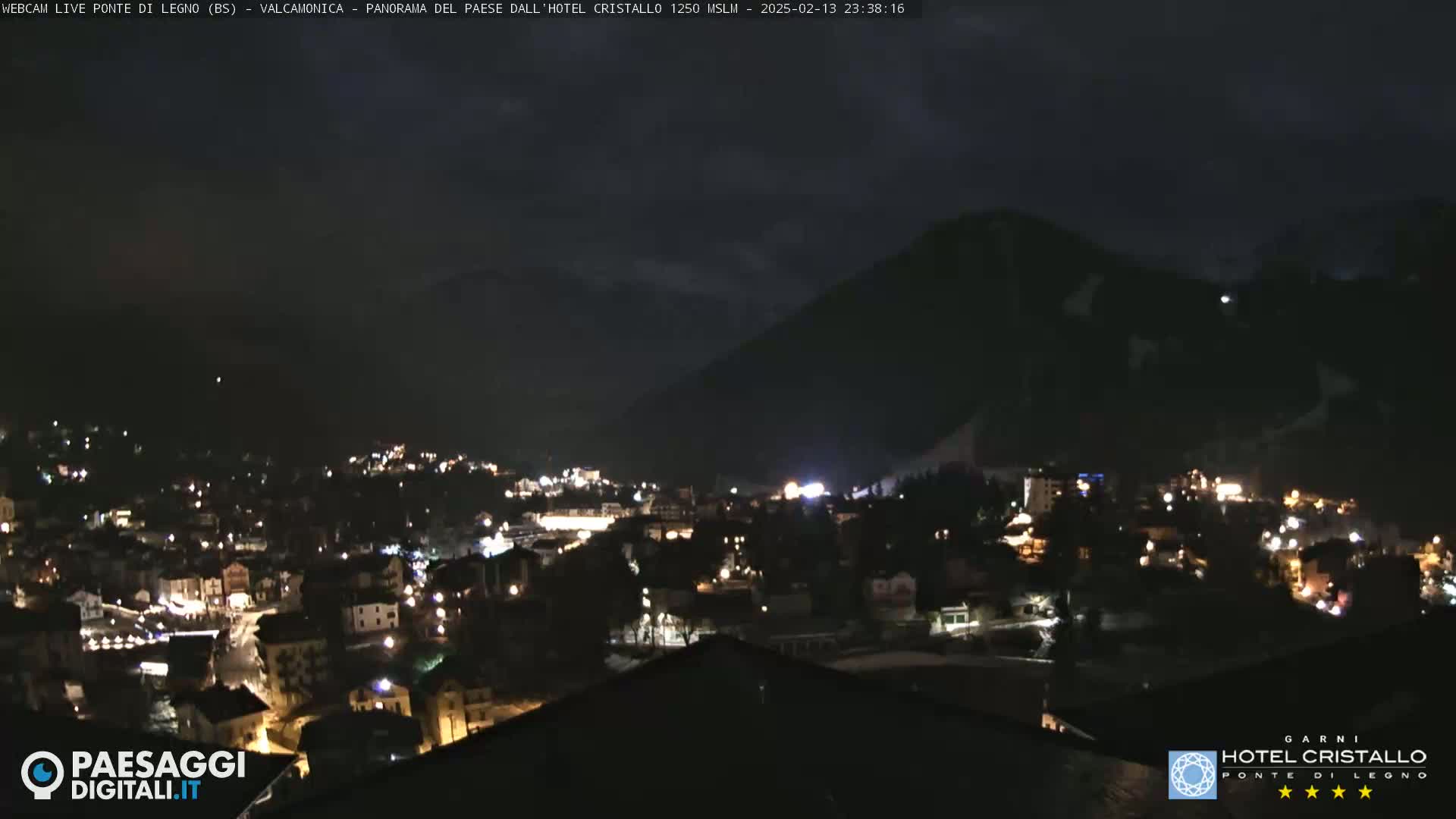 A nighttime view of a town nestled in a valley, illuminated by streetlights and buildings, with dark mountains in the background under a cloudy sky.