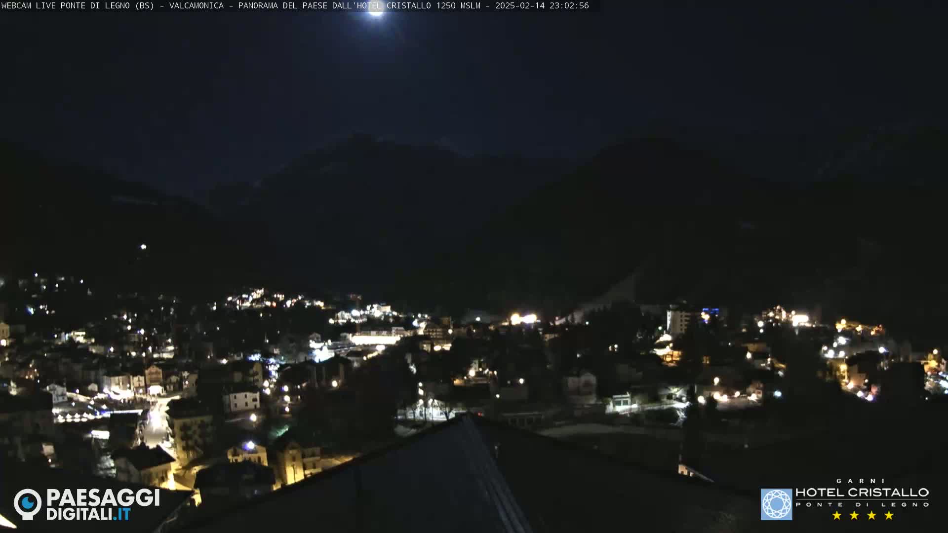 A nighttime view of a town nestled in a valley, illuminated by scattered lights, with mountains visible in the background under a dark, clear sky.
