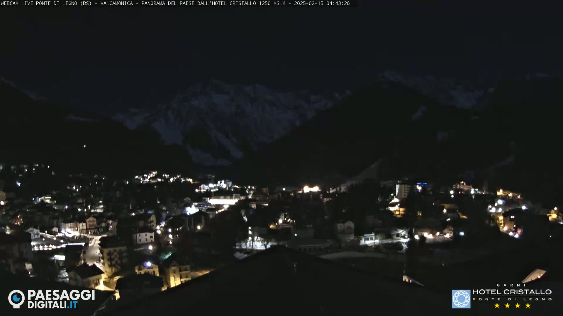 A nighttime view of a town nestled in a valley, illuminated by scattered lights, with dark mountains in the background under a clear, dark sky.