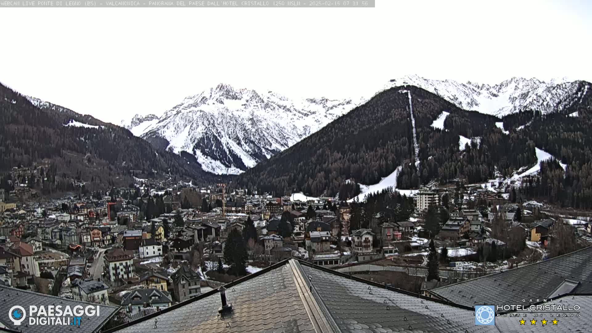 A snow-dusted mountain range overlooks a town nestled in a valley under a cloudy sky.