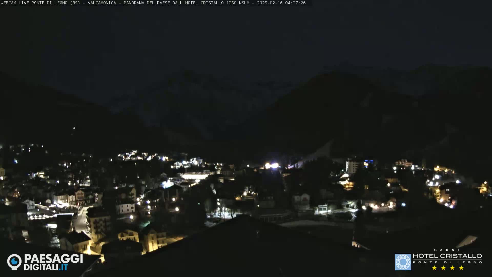 A nighttime view of a town nestled in a valley, illuminated by scattered lights, with dark mountains rising in the background under a clear, dark sky.