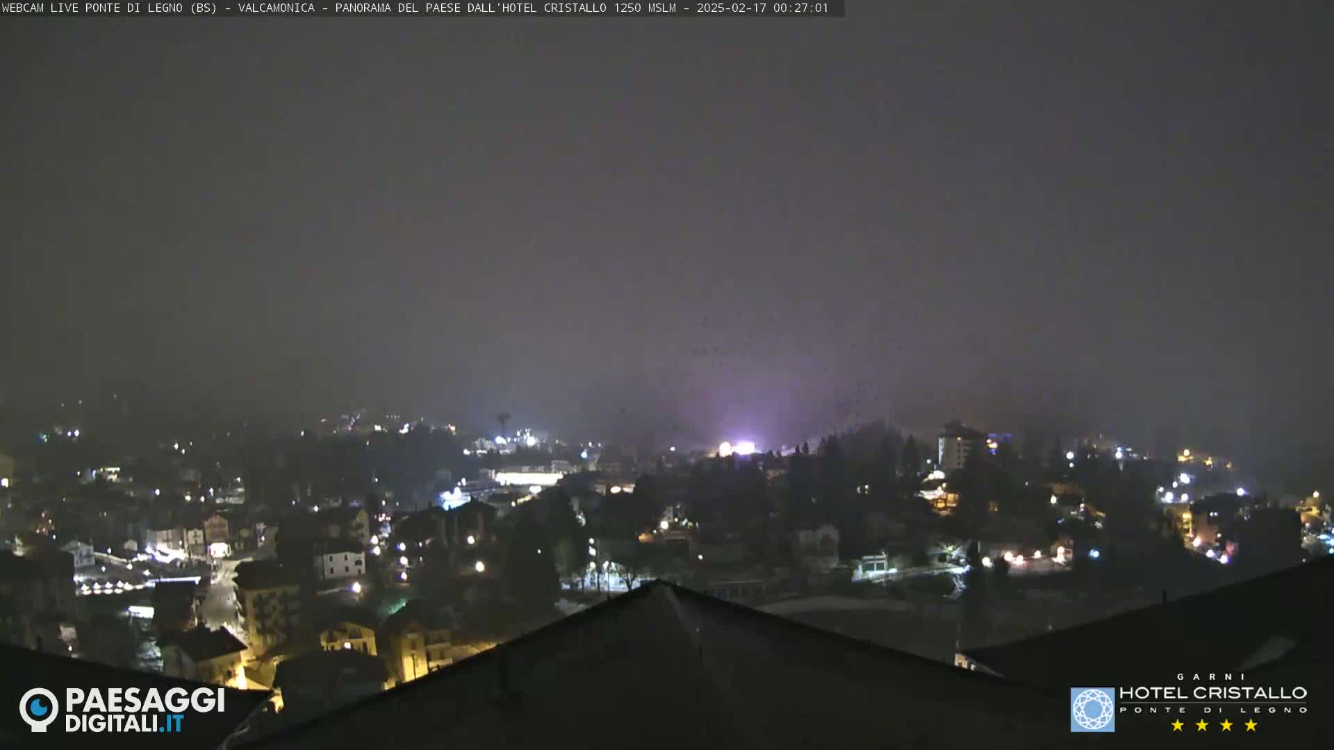 A nighttime, hazy view of a town nestled in a valley, with scattered lights visible under a dark, possibly snowy sky.