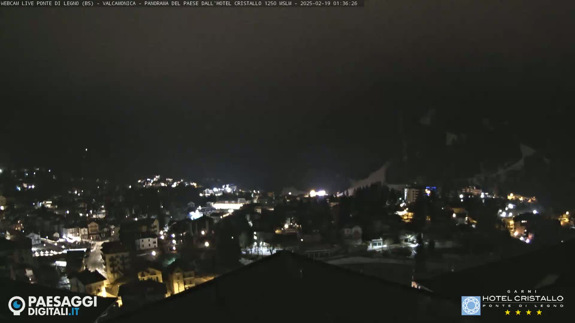 A nighttime view of a town nestled in a valley, with sparsely lit buildings and a dark, possibly cloudy sky.