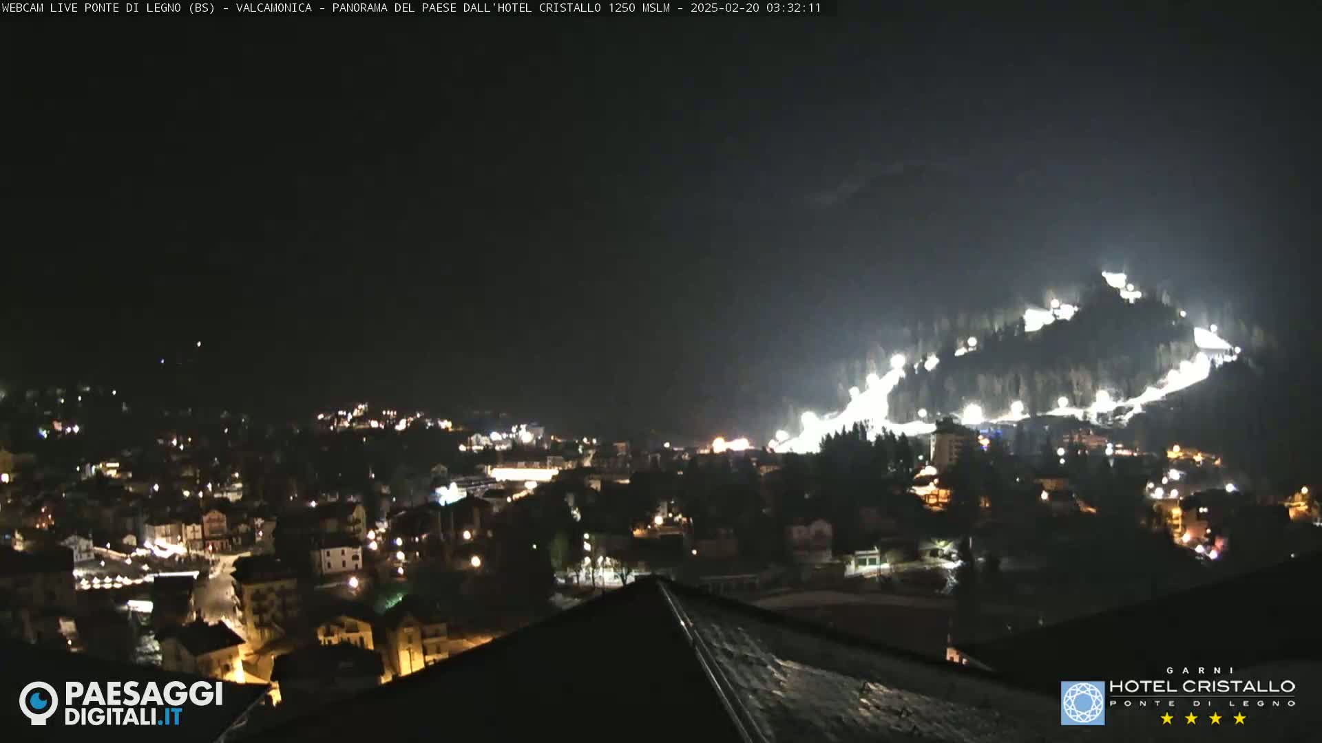 A nighttime view of a town nestled in a valley, illuminated by lights, with a brightly lit ski slope on a hillside in the background under a dark sky.
