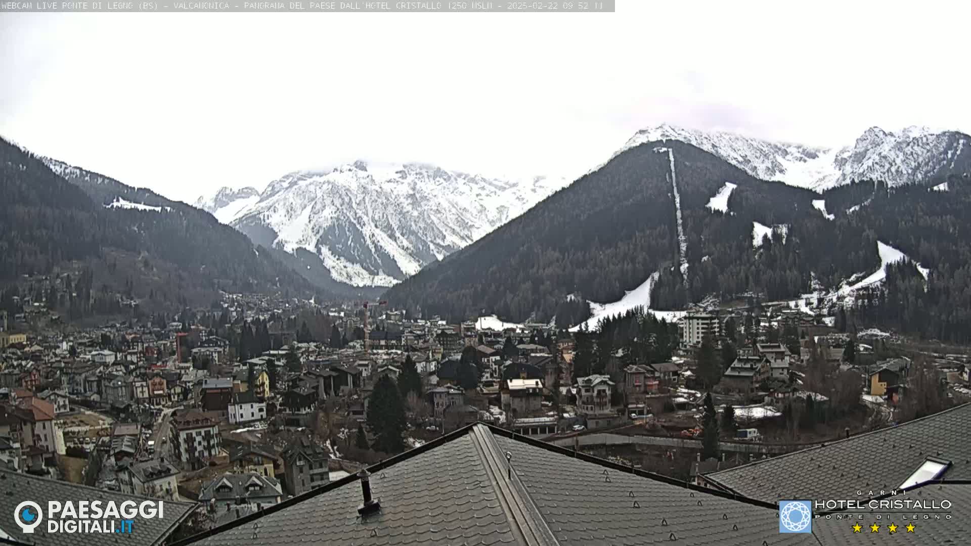 A view from a rooftop shows a town nestled in a valley between snow-capped mountains on an overcast day.