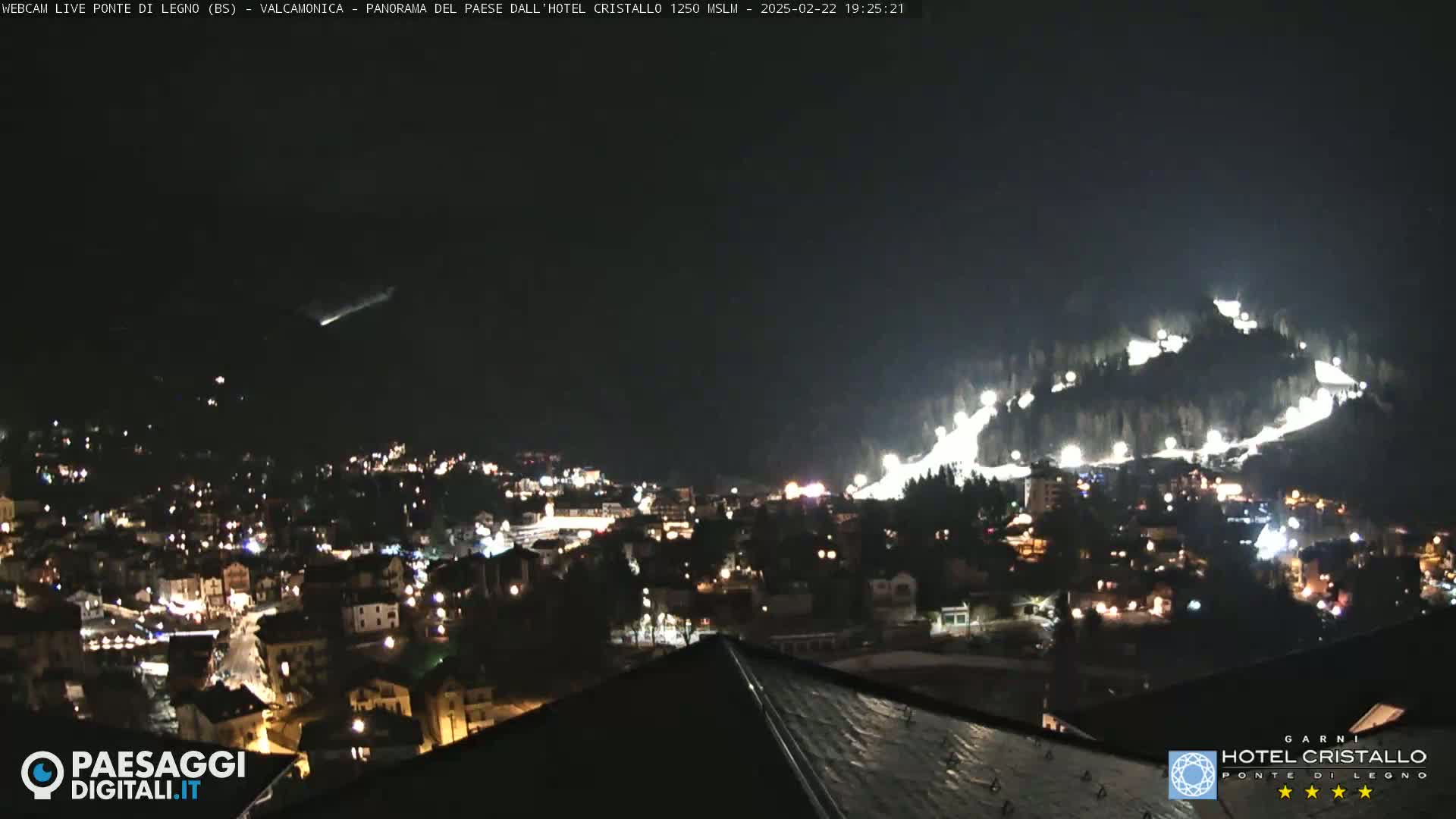 A nighttime view of a snow-covered hillside with illuminated ski slopes overlooking a town with many lights.