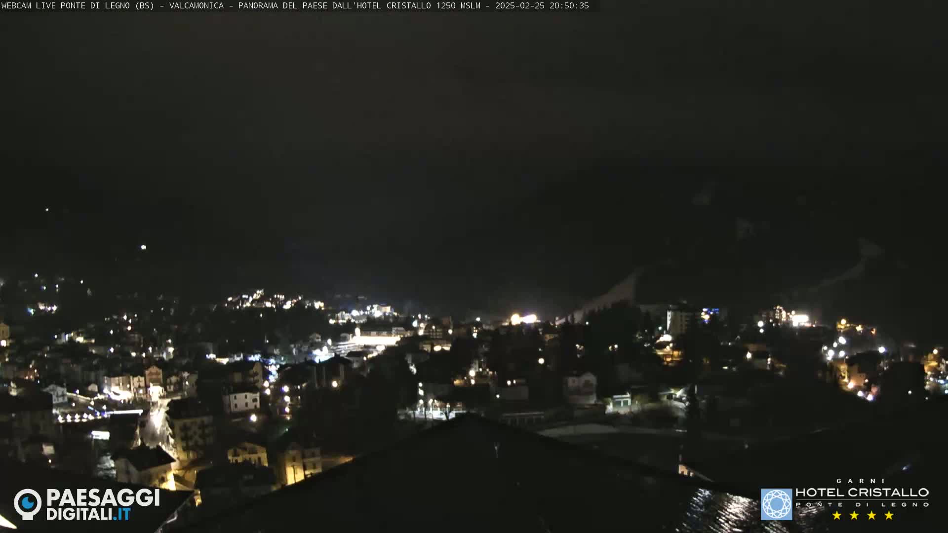 A nighttime view of a town illuminated by lights, nestled in a valley under a dark, possibly cloudy sky.
