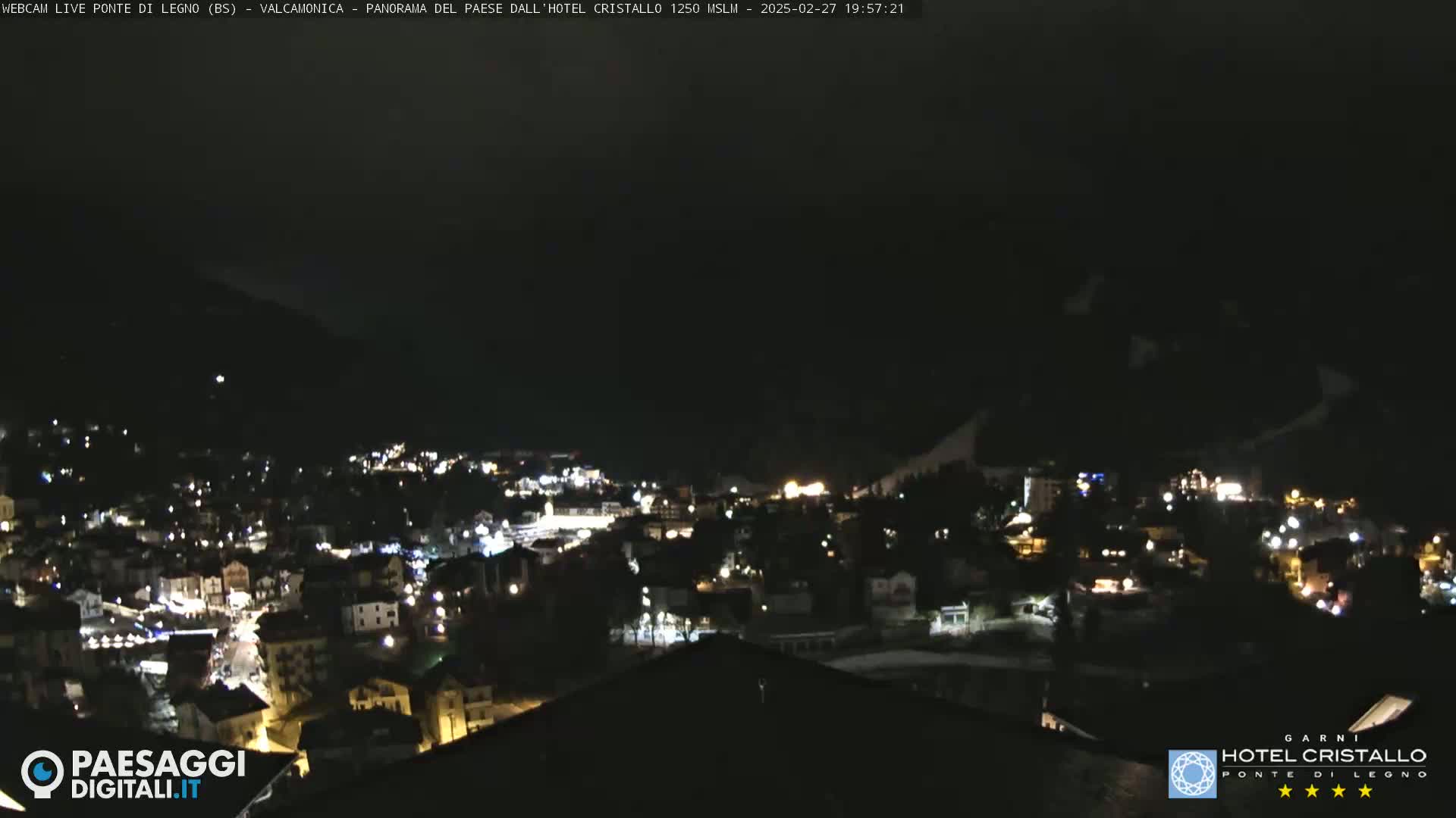 A nighttime view of a town nestled in a valley, illuminated by scattered lights under a dark sky.