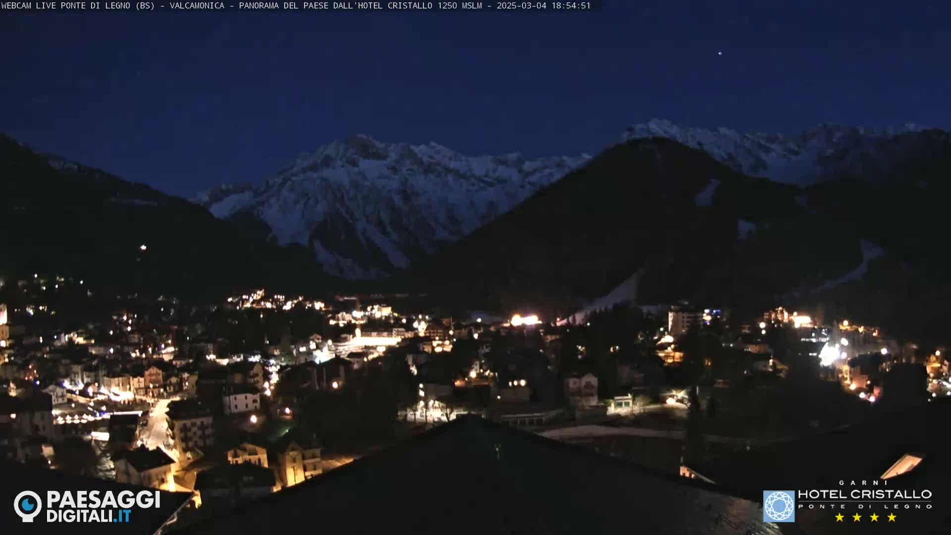 A nighttime view of a snow-capped mountain range overlooking a small, illuminated town under a clear, dark sky.