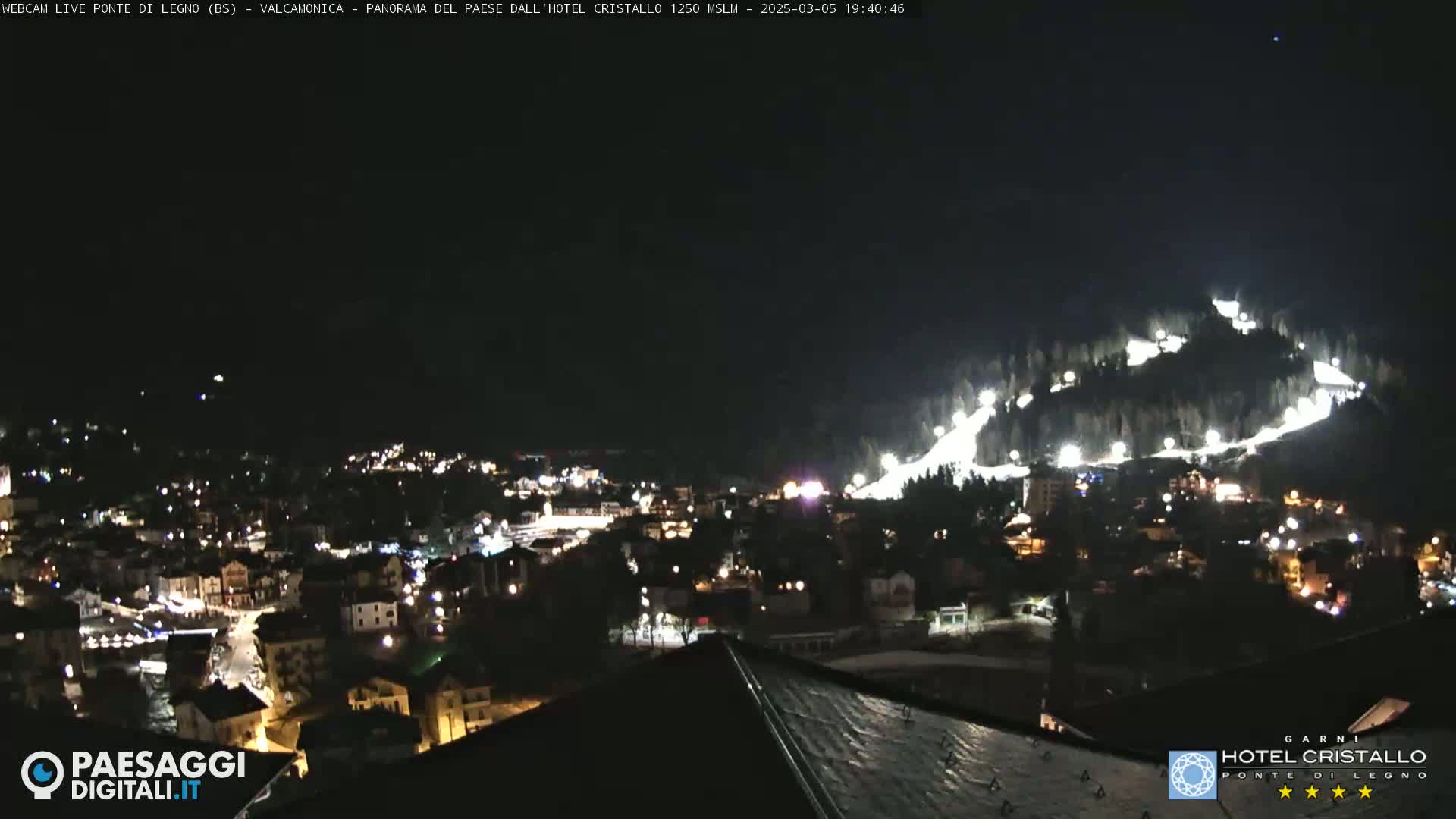A nighttime view of a town nestled in a valley, with illuminated ski slopes on a hillside above.