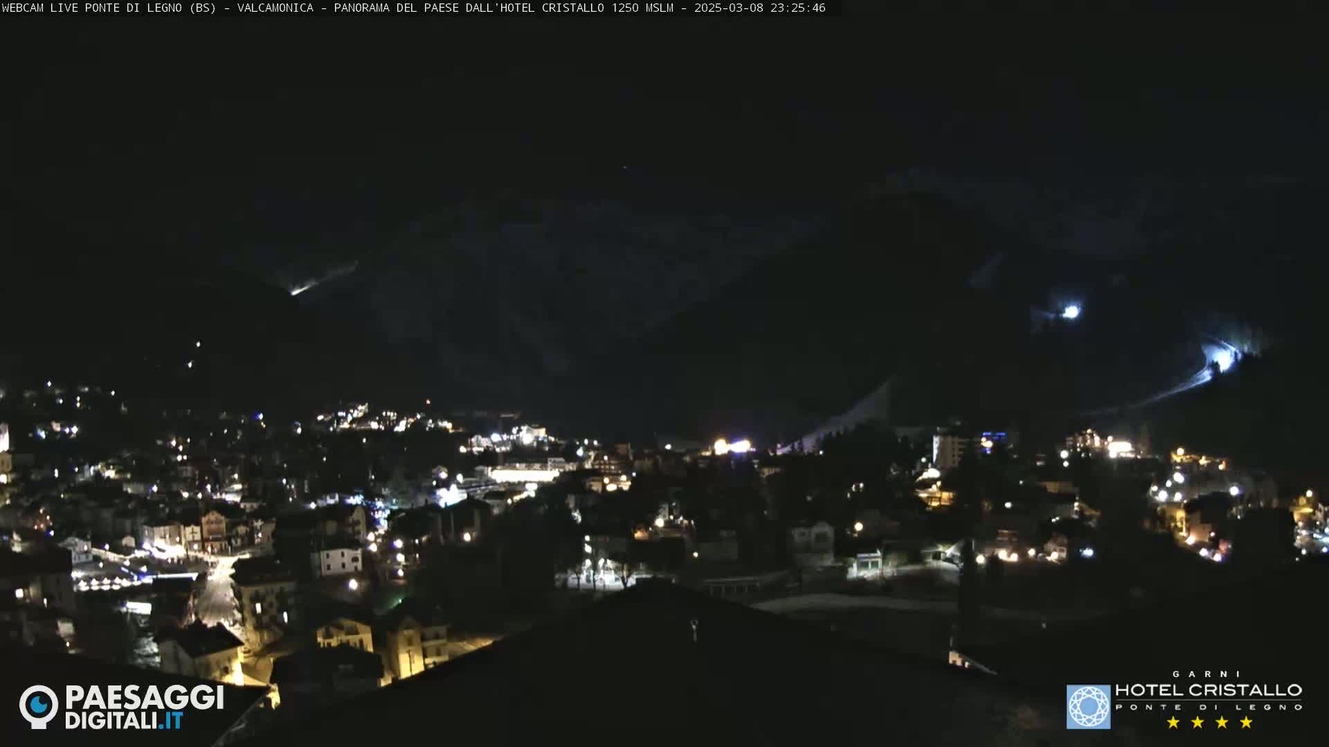 A nighttime view of a town nestled in a valley, illuminated by lights, with snow-covered mountains in the background under a clear sky.