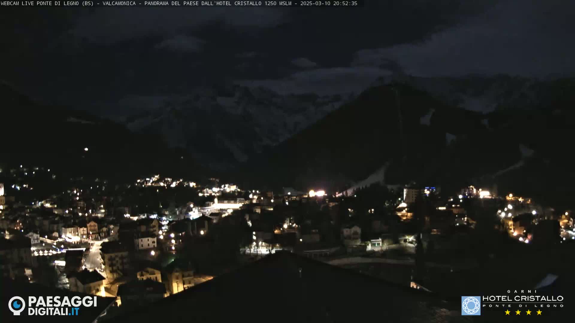 A nighttime view of a town nestled in a valley, illuminated by streetlights, with snow-covered mountains in the background under a cloudy sky.