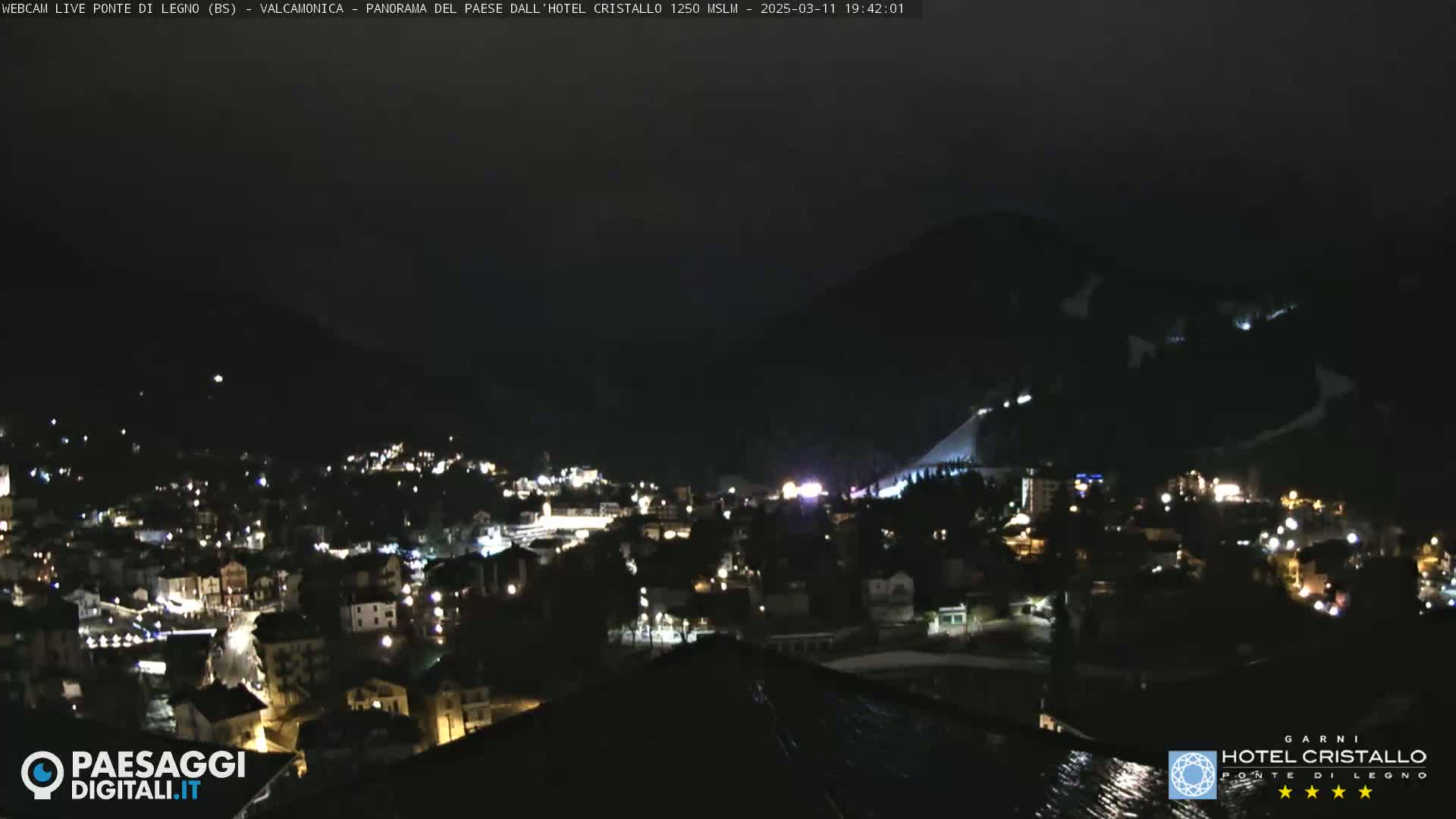 A nighttime view of a town nestled in a valley beneath a dark mountain, illuminated by scattered lights.