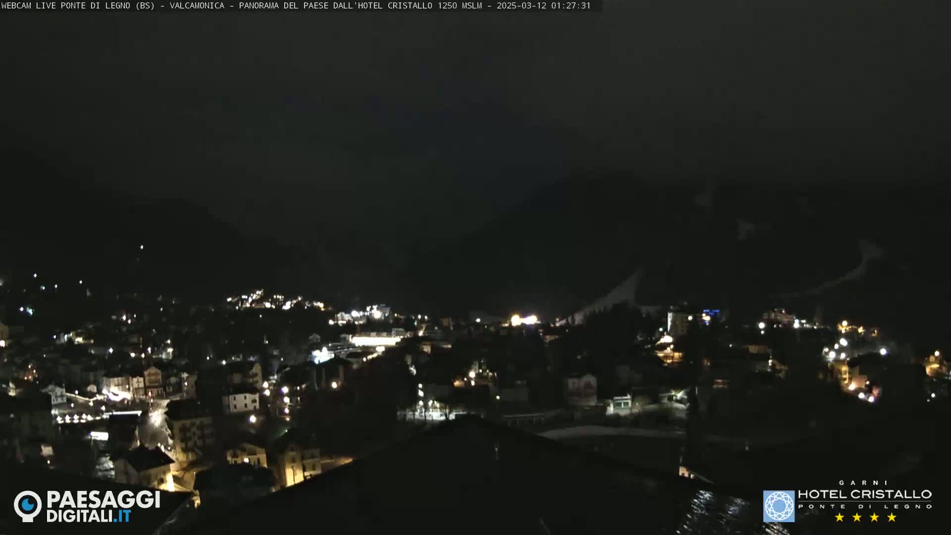A nighttime view of a town nestled in a valley, illuminated by scattered lights against a dark mountain backdrop under a clear, dark sky.