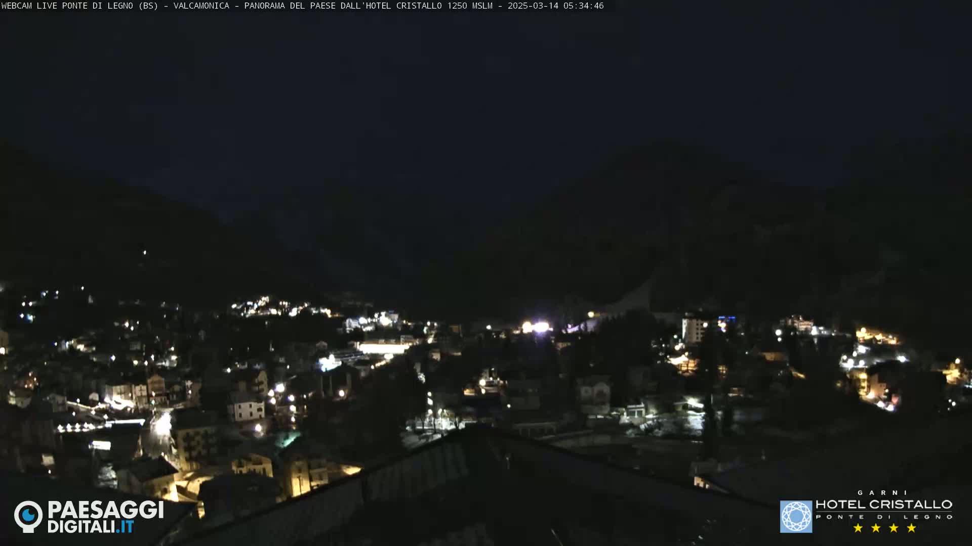 A nighttime view of a town nestled in a valley, illuminated by scattered lights under a dark sky.