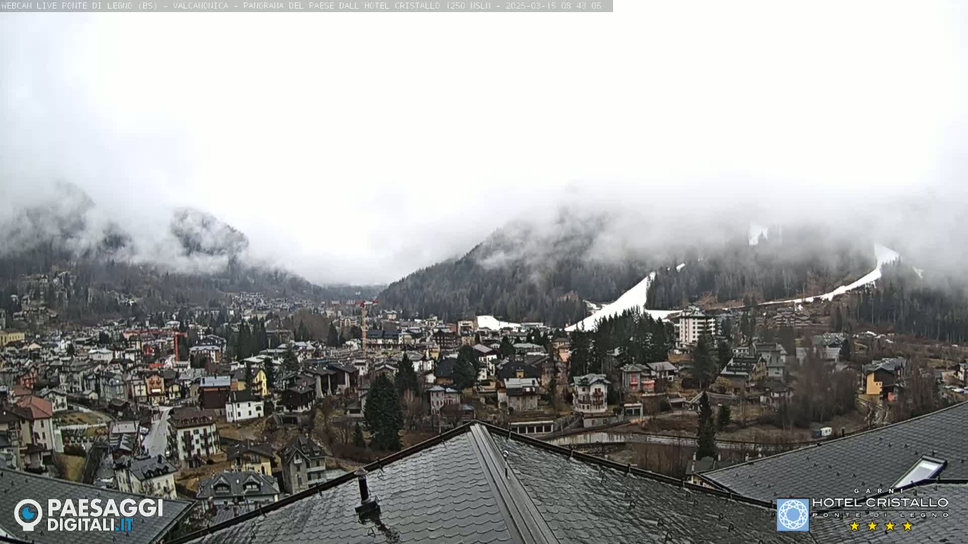 A misty mountain town sits below fog-covered hills, partially snow-capped, with a wet rooftop in the foreground.