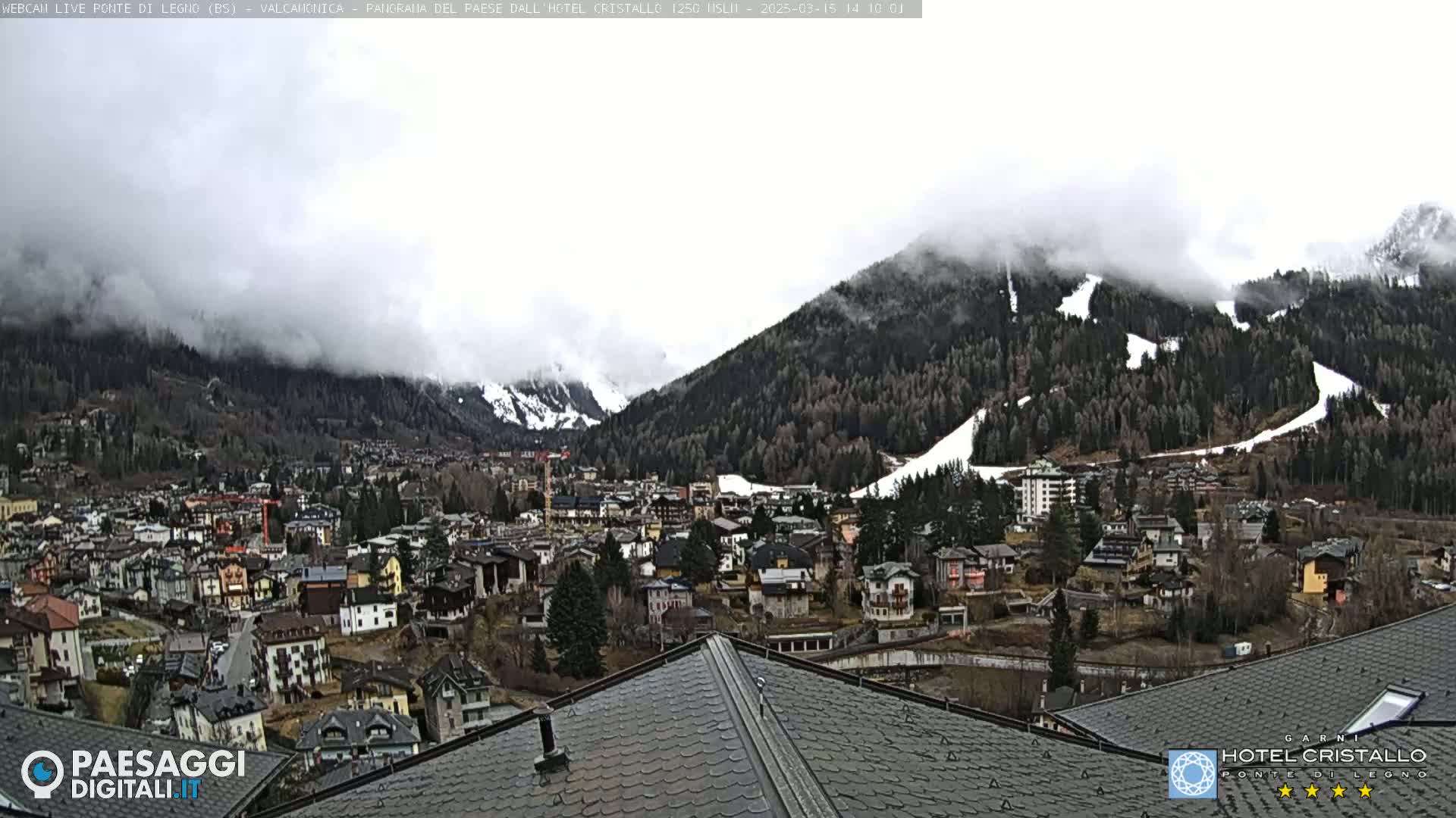 A mountain town nestled in a valley, partially obscured by fog and low clouds, with snow-covered mountain slopes in the background.