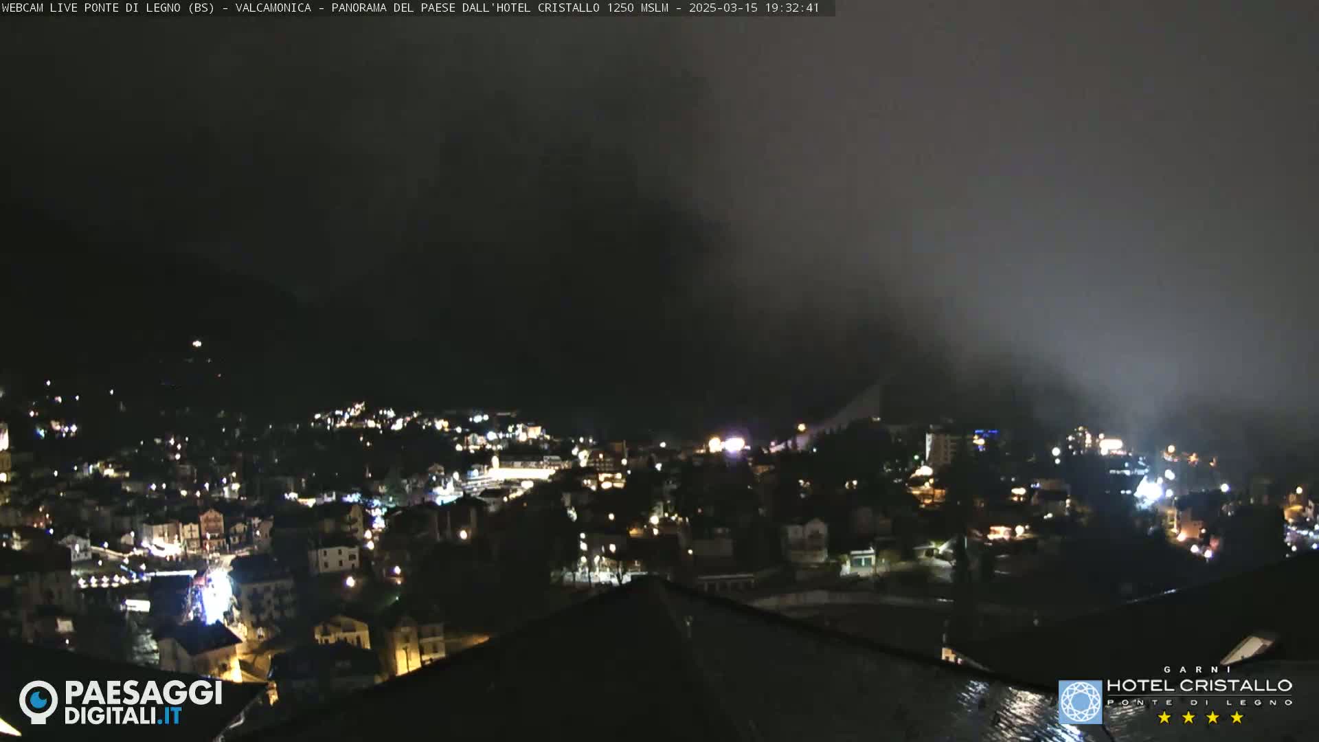 A nighttime view of a town nestled in a valley, partially obscured by fog, with the lights of buildings illuminating the scene.