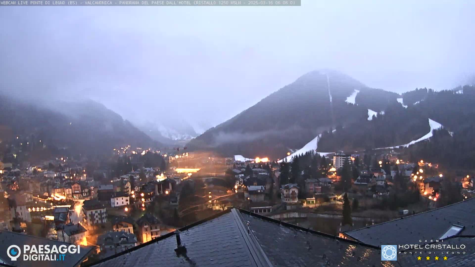 A nighttime view of a town nestled in a valley between snow-covered mountains under a cloudy sky.