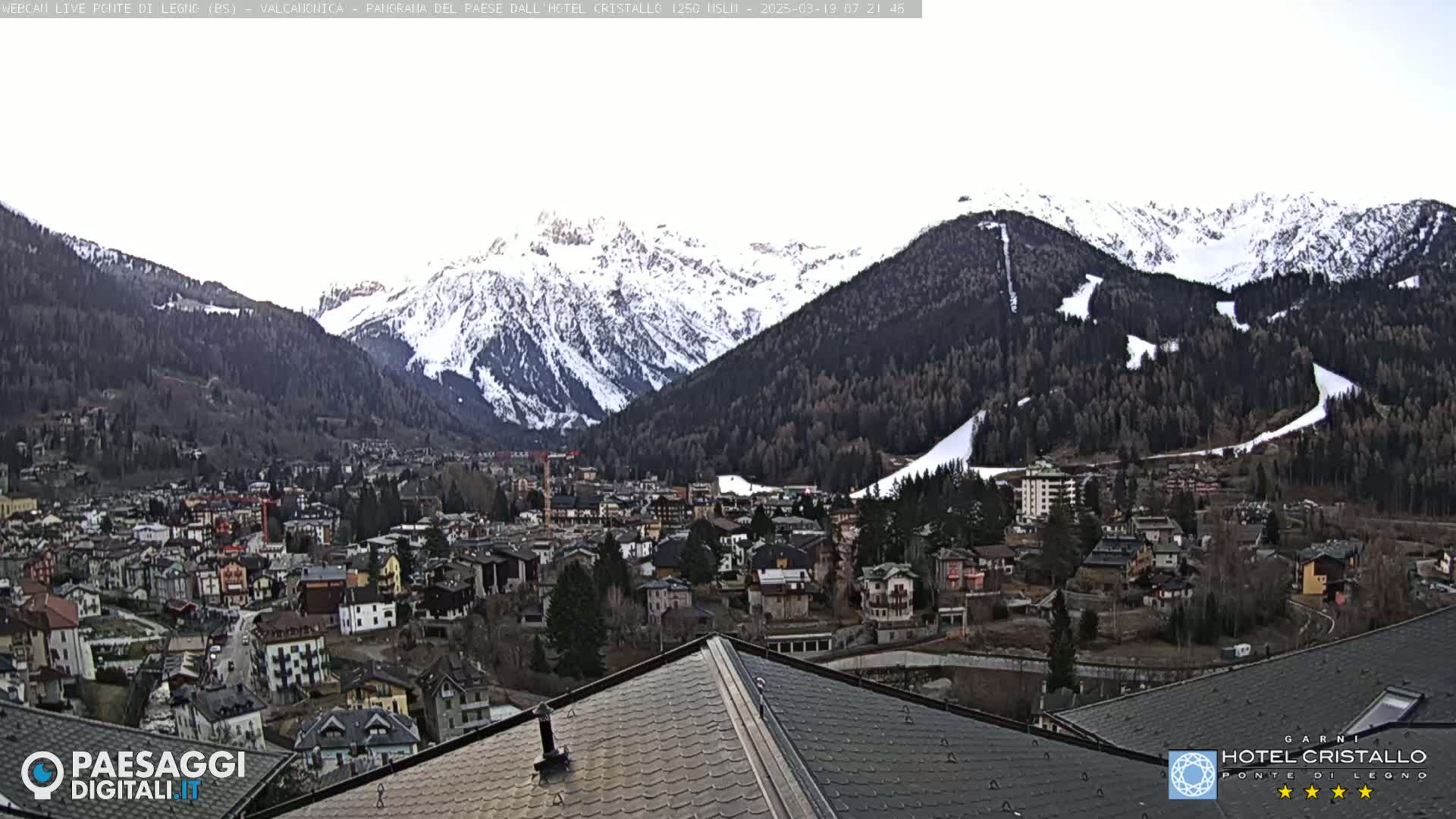 A mountain town nestled in a valley, with snow-capped mountains in the background under an overcast sky.