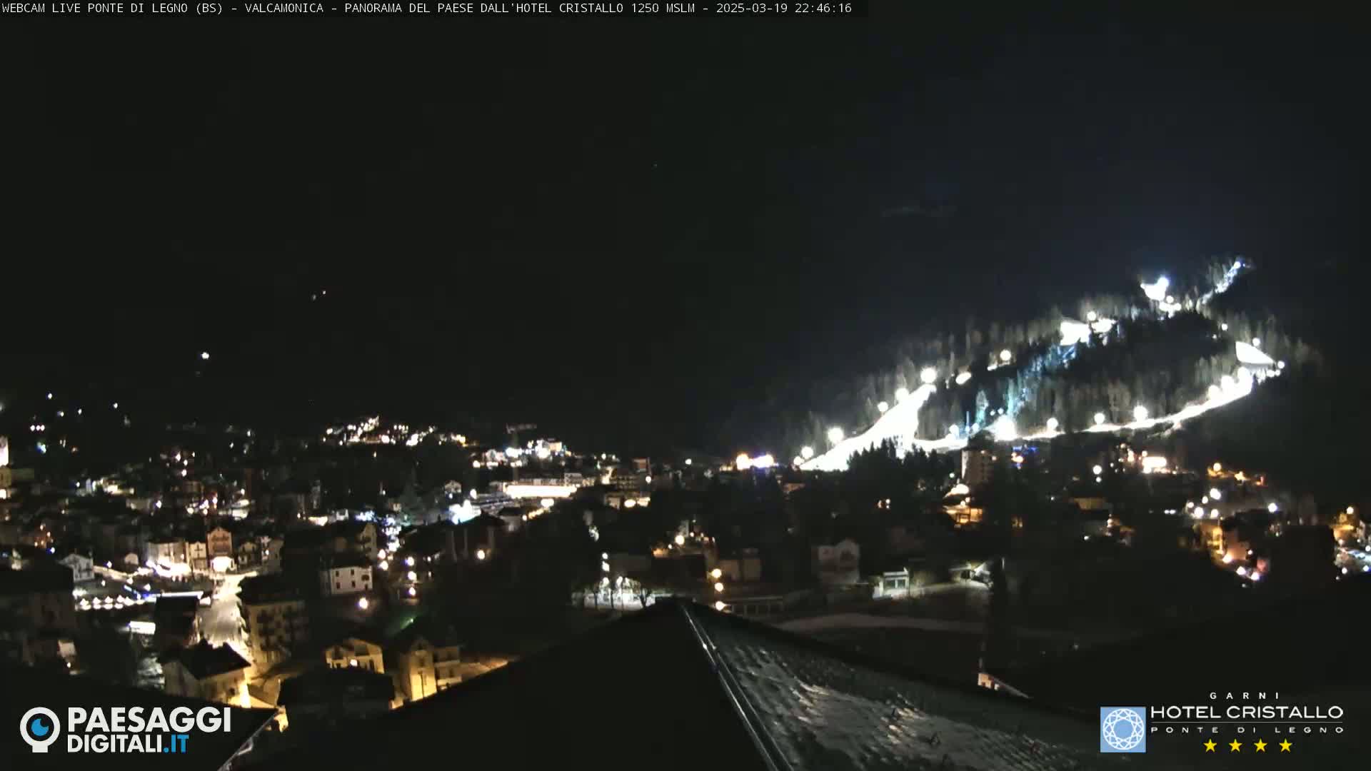 A nighttime view of a town nestled in a valley, with brightly lit ski slopes visible on a hillside above.