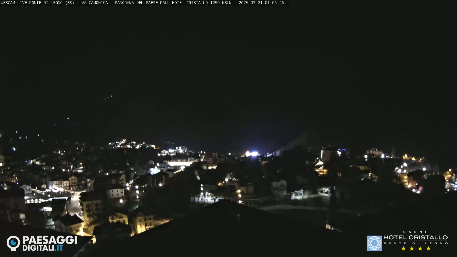 A nighttime, high-angle view shows a town nestled in a valley, illuminated by scattered lights under a clear, dark sky.